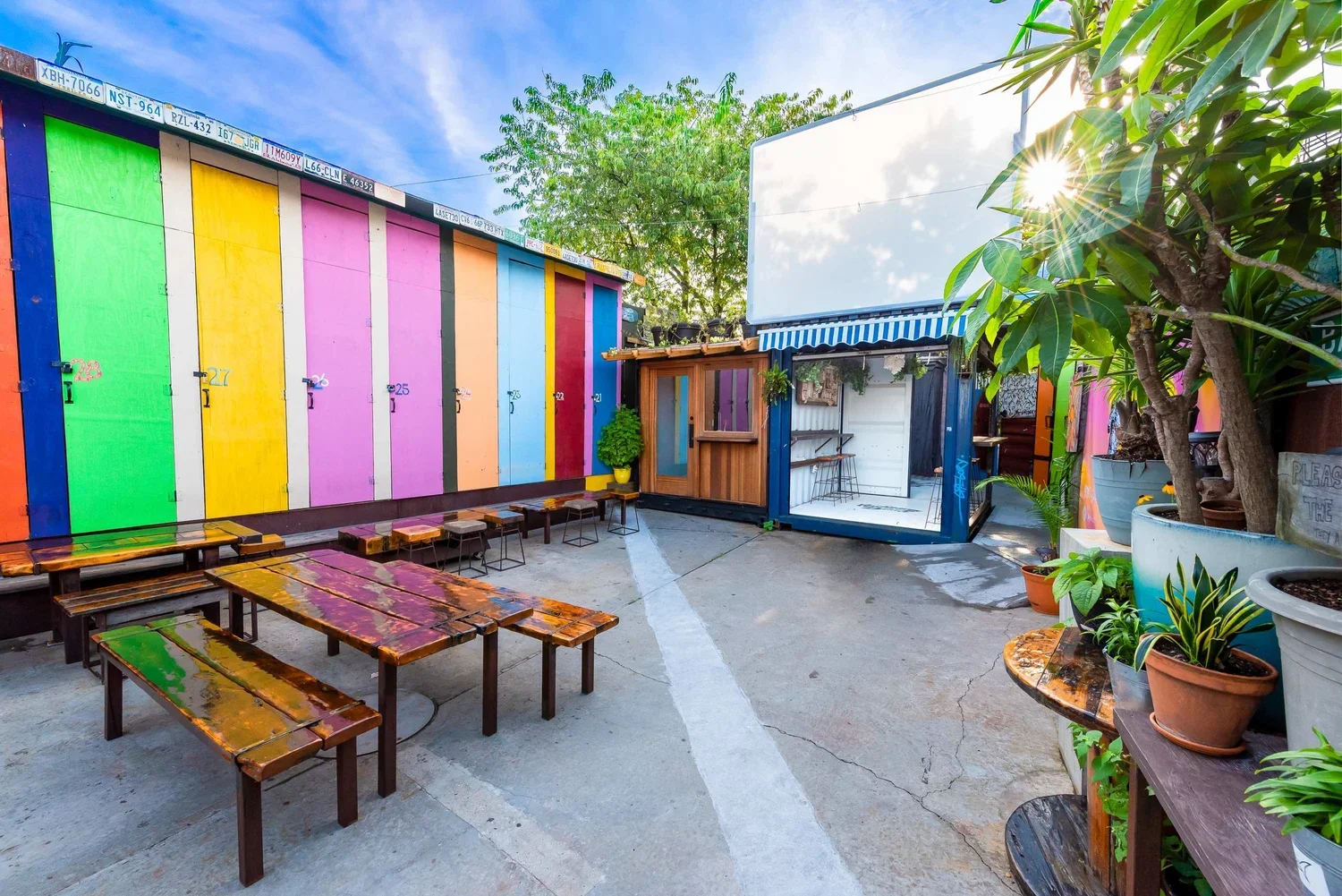 Colorful outdoor patio with painted wooden surf lockers, wooden benches, potted plants, and a small wooden structure with a striped awning, on a sunny day.