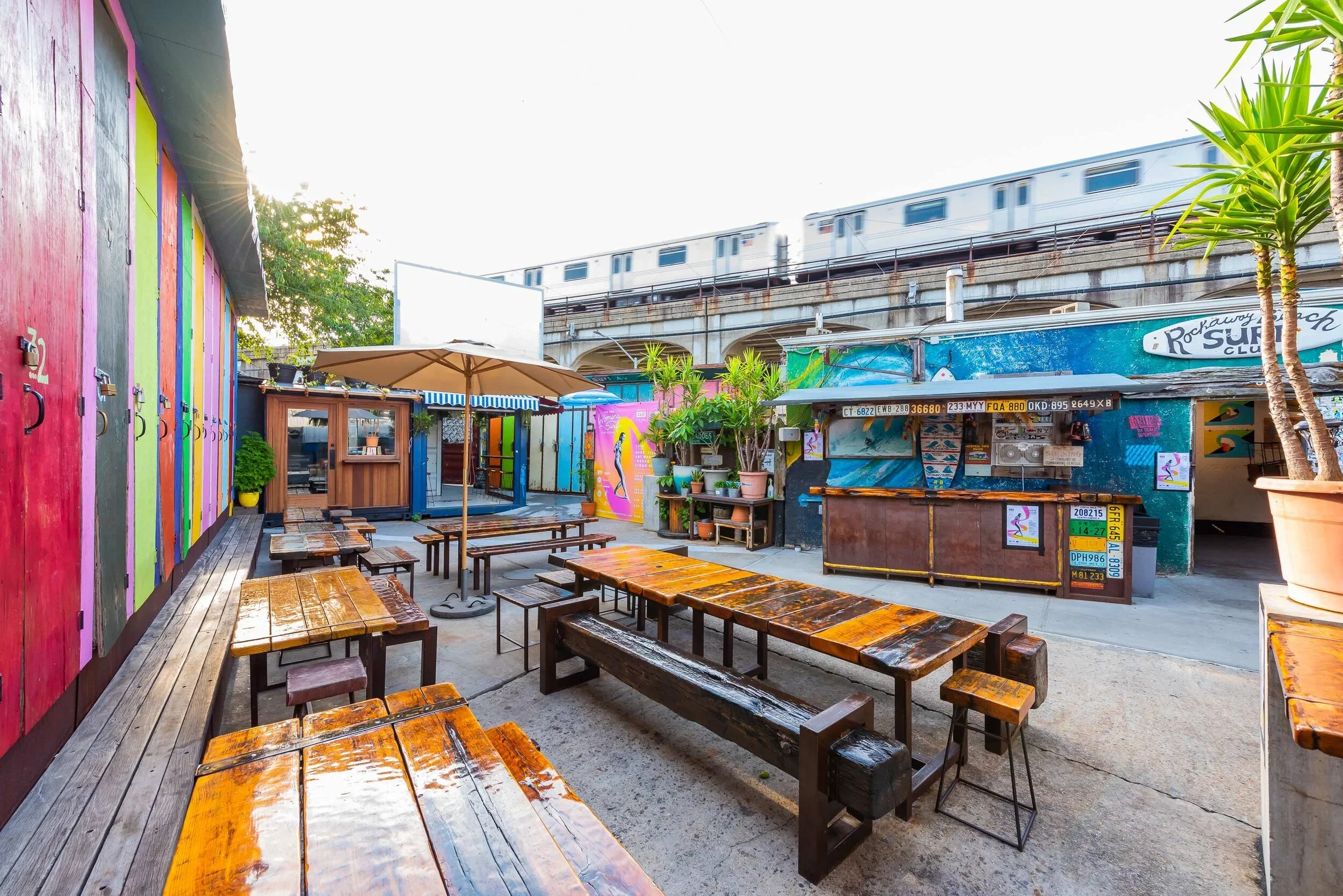 Outdoor seating area with wooden tables and benches, colorful painted walls, potted plants, and a small counter under an umbrella, with train tracks and train passing overhead in the background.