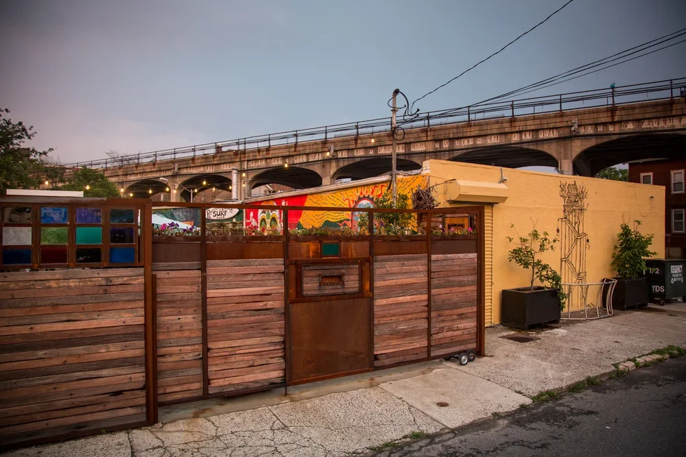 Exterior view of Rockaway Beach Surf Club shown with a mural painted on its side, surrounded by planters and a wooden fence, with an elevated subway in the background at dusk.