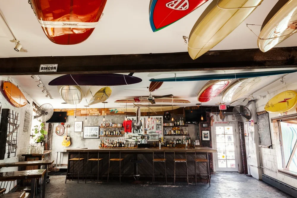 Interior of a bar with surfboards hanging from the ceiling and a variety of bottles behind the bar counter.