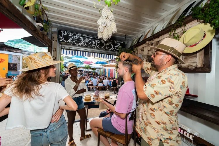 A man gets a hat braided by a woman in a pink shirt at an outdoor market or event, with other people and umbrellas visible in the background.