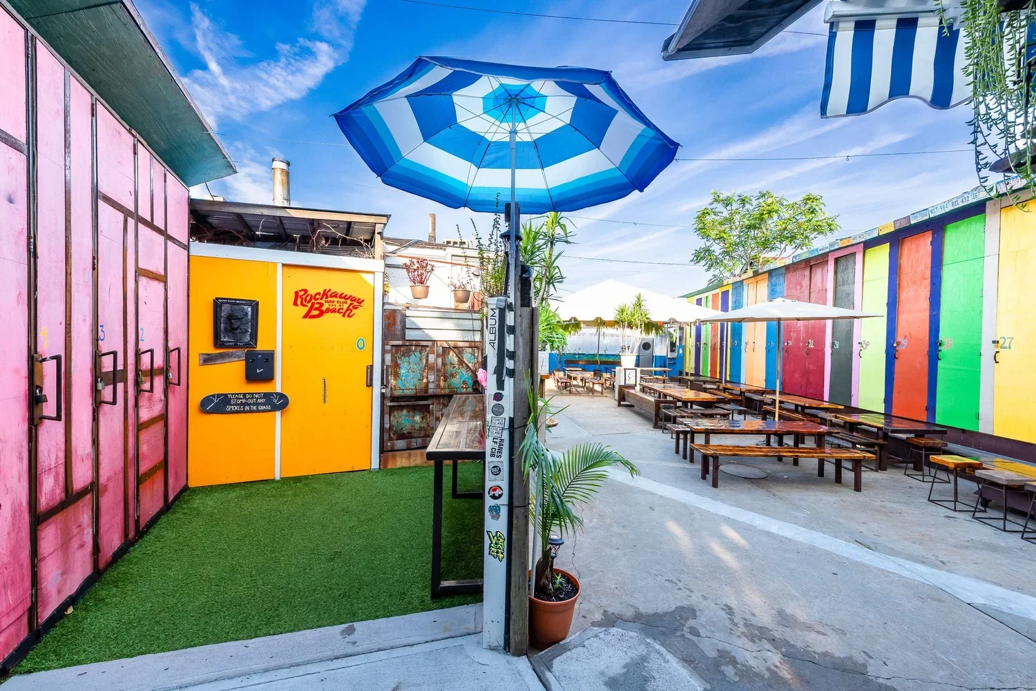 Colorful outdoor patio with rainbow-colored lockers on the right, picnic tables with umbrellas, a yellow wall with 'Rockaway Beach' sign, a blue and white striped umbrella overhead, and blue skies with clouds.