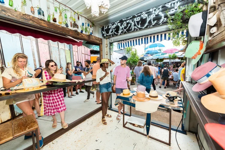 People browsing hats at a market stall with more shoppers in the background, umbrellas providing shade, and colorful decor on the walls.