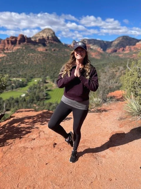A woman stands on a desert trail in front of red rock formations and mountains, smiling with hands in a prayer position, wearing workout clothes and a hat on a sunny day.