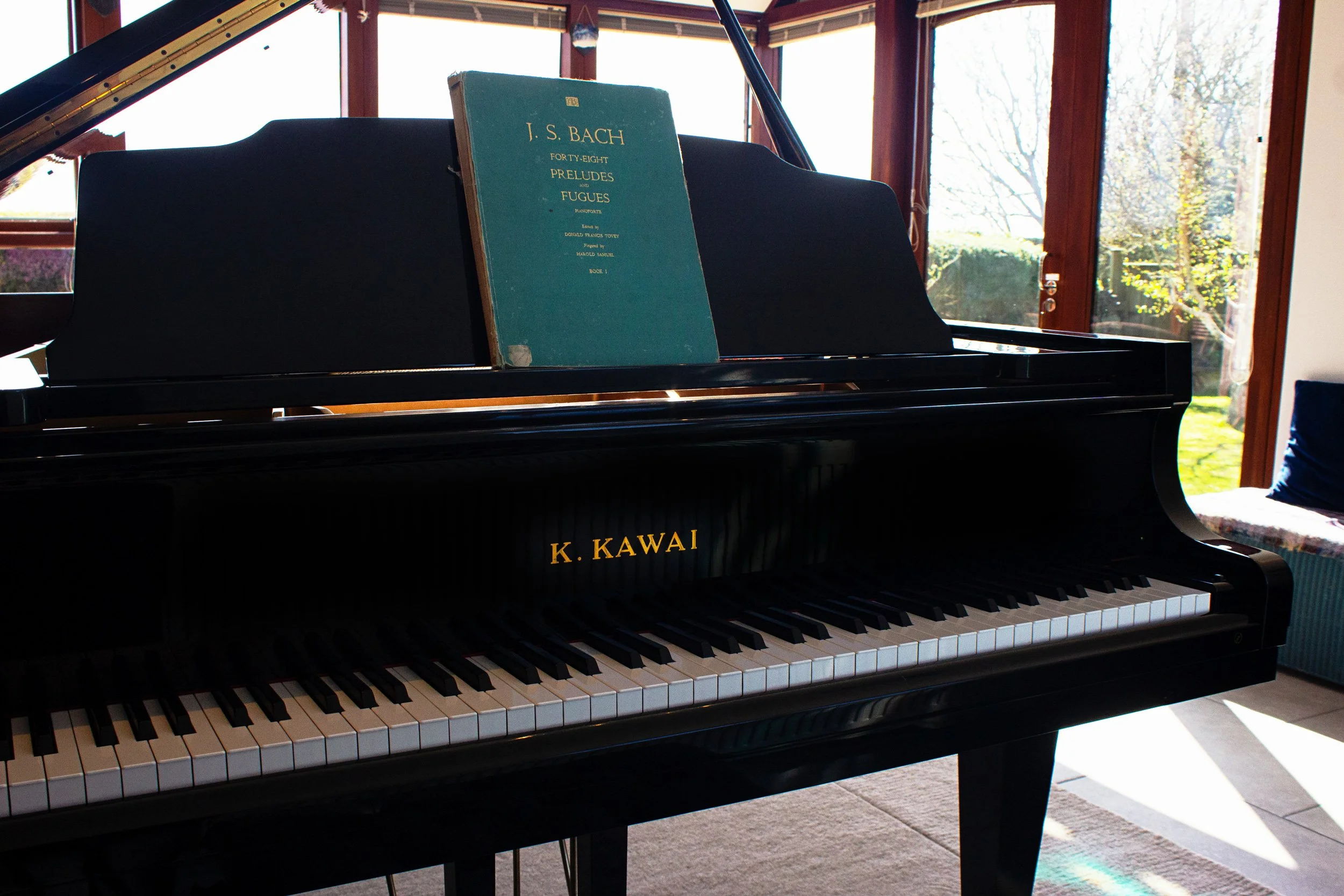 A black grand piano with a sheet of music titled 'J. S. Bach Forty-Eight Preludes and Fugues' placed on the music stand, situated in a room with large windows and natural daylight.