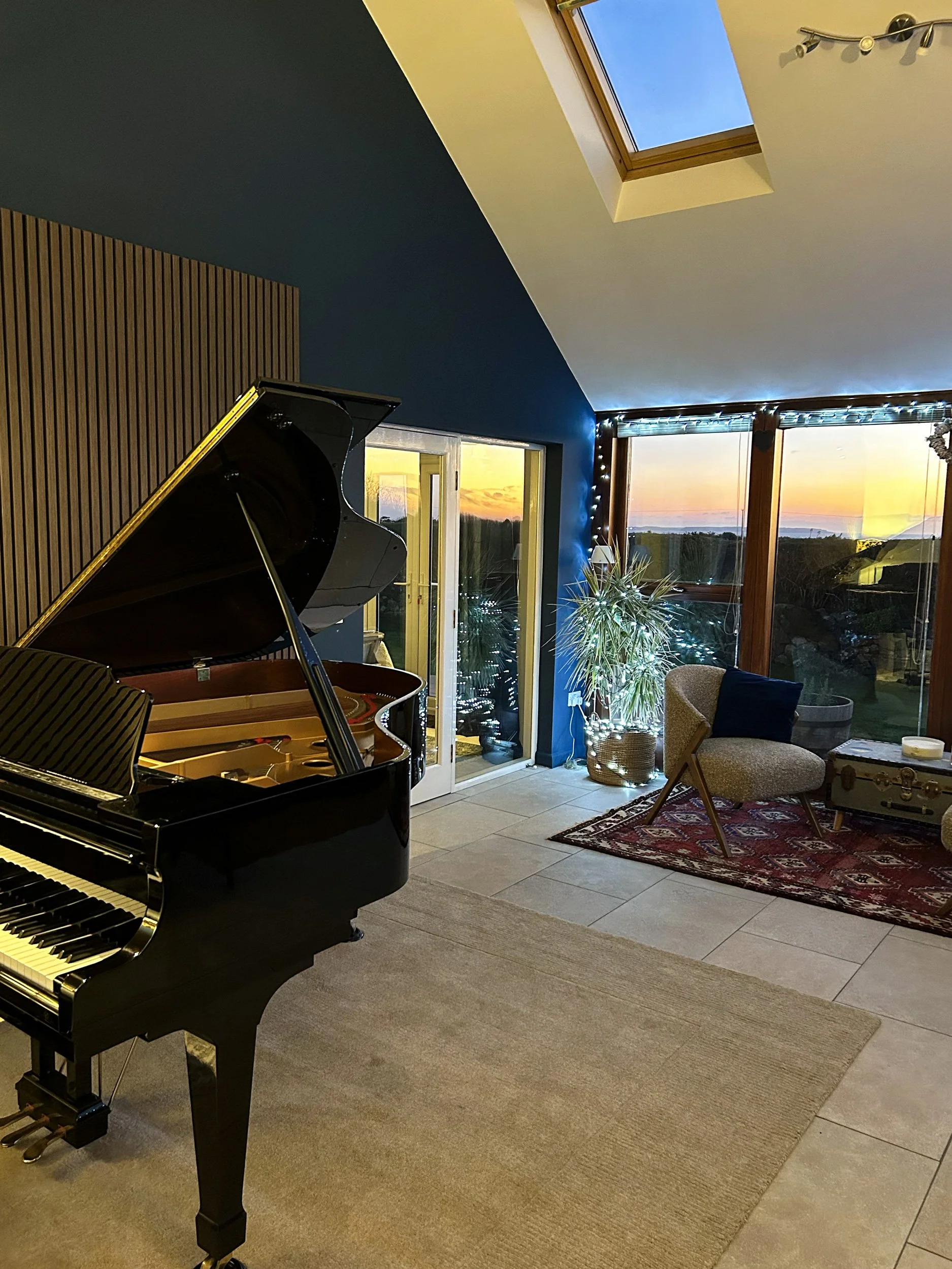 The Music Room St Davids with a black grand piano, a beige armchair with a dark pillow, a small side table, and large windows showing a sunset sky. There is a rectangular rug and a potted plant next to the window, with ceiling lights and a skylight.