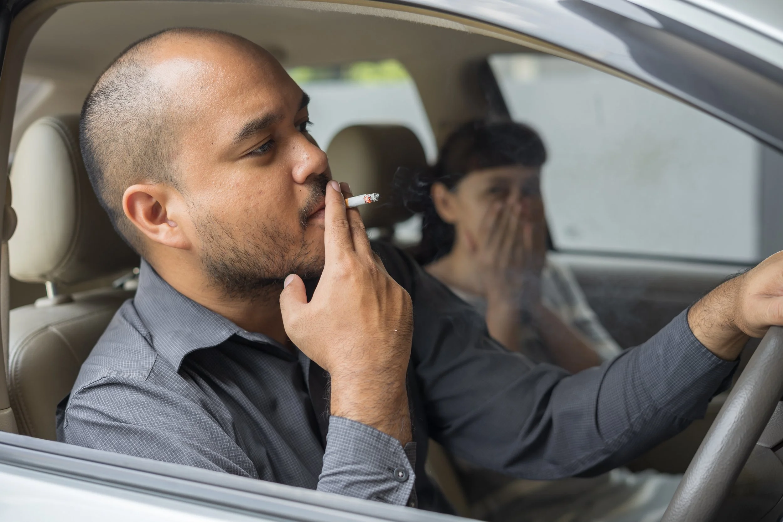 A man and a woman sitting in a car, with the man smoking a cigarette and the woman covering her mouth, possibly appearing upset or coughing.