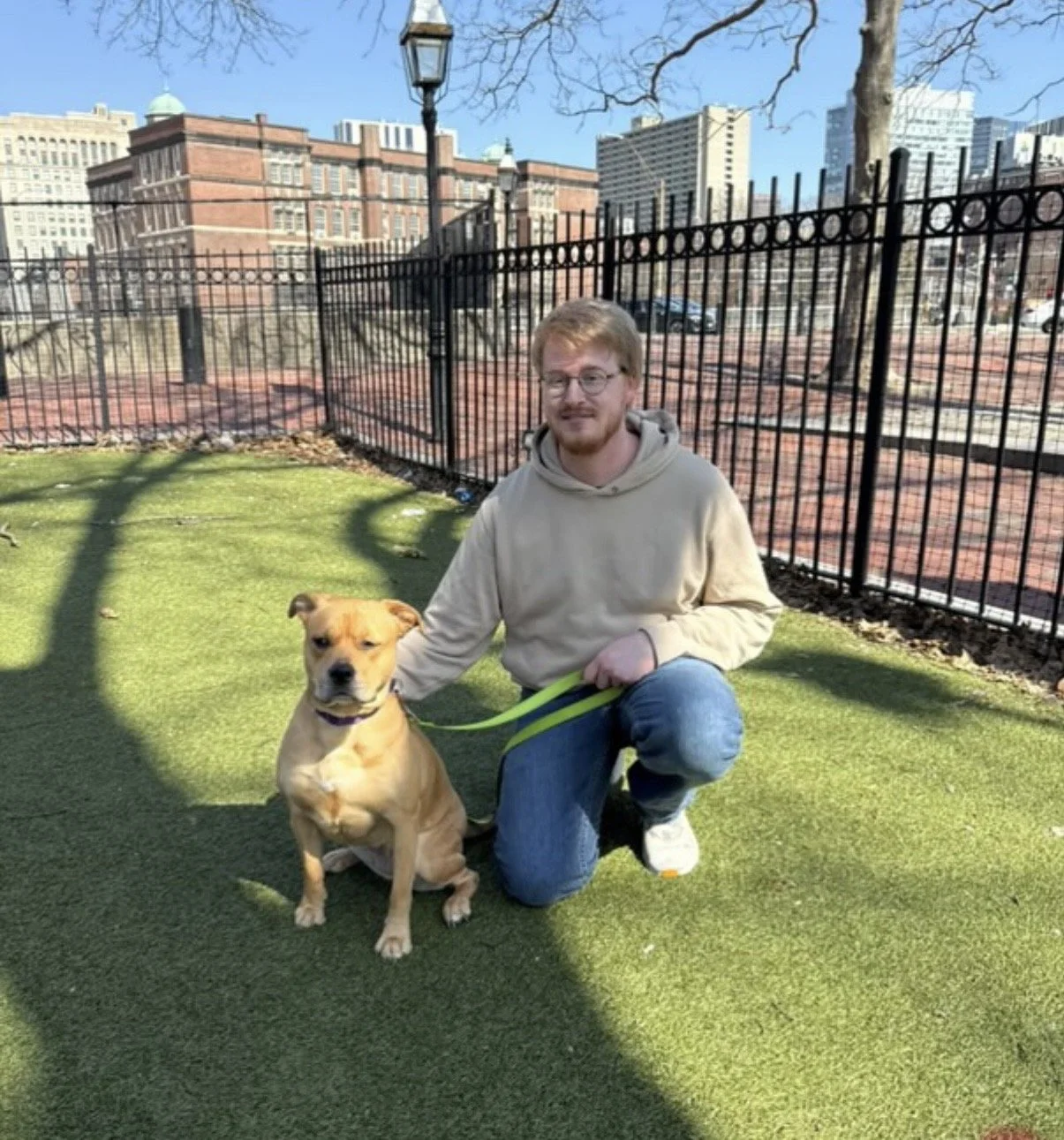 A man crouching on green grass holding a tan dog on a leash in a dog park and city buildings in the background on a sunny day.