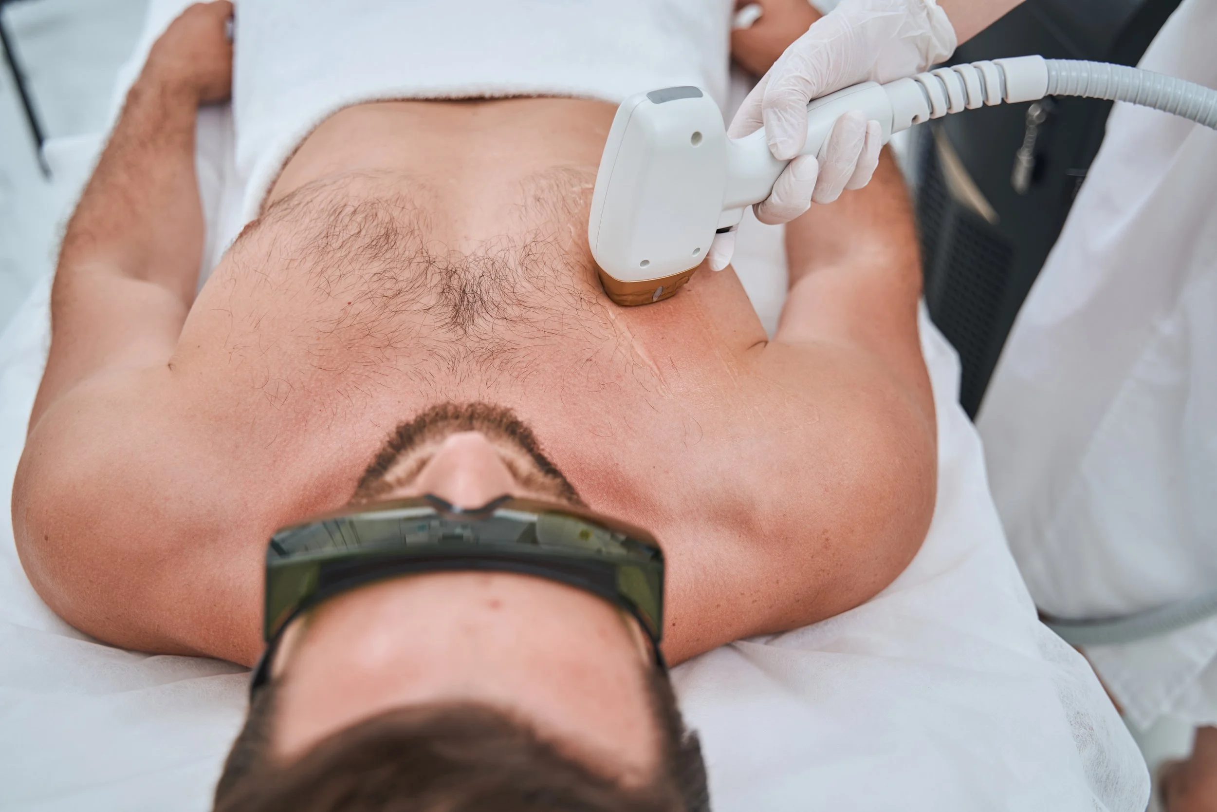 En person ligger ned på en legeplass, blir analysert med en maskin av en helsepersonell med hvite hansker. Personen har på seg briller og kjortel, og ligger på en hvit seng.
