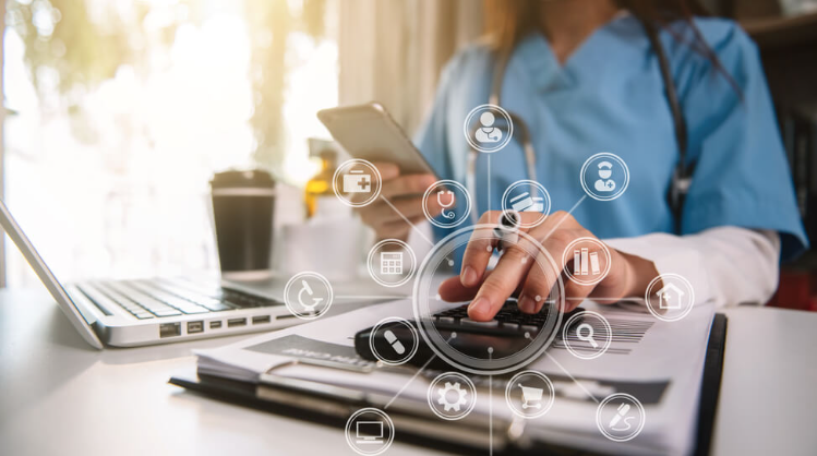 A healthcare professional in scrubs using a tablet with digital medical icons floating around, beside a laptop and paperwork on a desk.