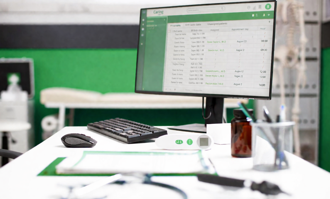 Medical office desk with computer monitor displaying patient schedule, keyboard, mouse, brown pill bottle, and various office supplies.