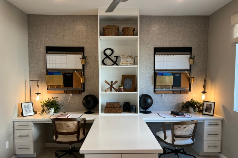 Home office with two desks, each with a chair and decorative items, flanking a central white bookshelf with baskets, decor, and electronics, backed by a textured wall and lit by wall-mounted lights.