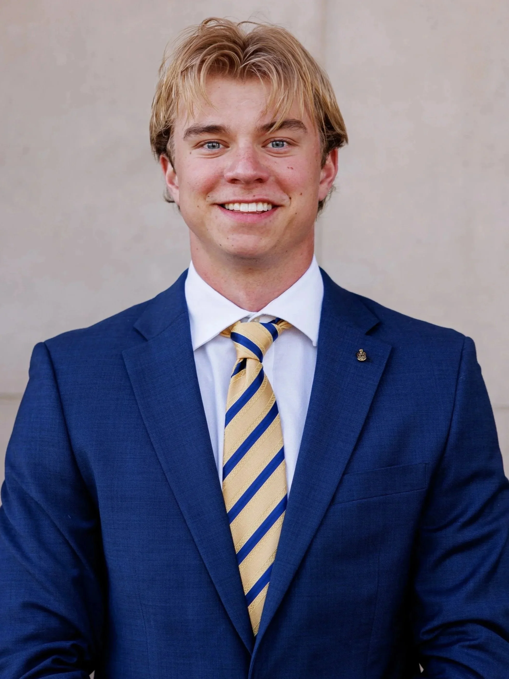 A young man with blonde hair, blue eyes, and a big smile, wearing a blue suit, white shirt, and yellow striped tie.