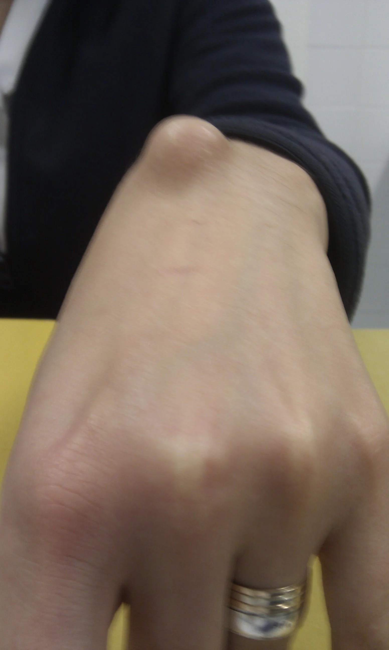 Close-up of an arm and shoulder of a person wearing a black long sleeve shirt, with jewelry on the wrist, touching a yellow surface in the background.
