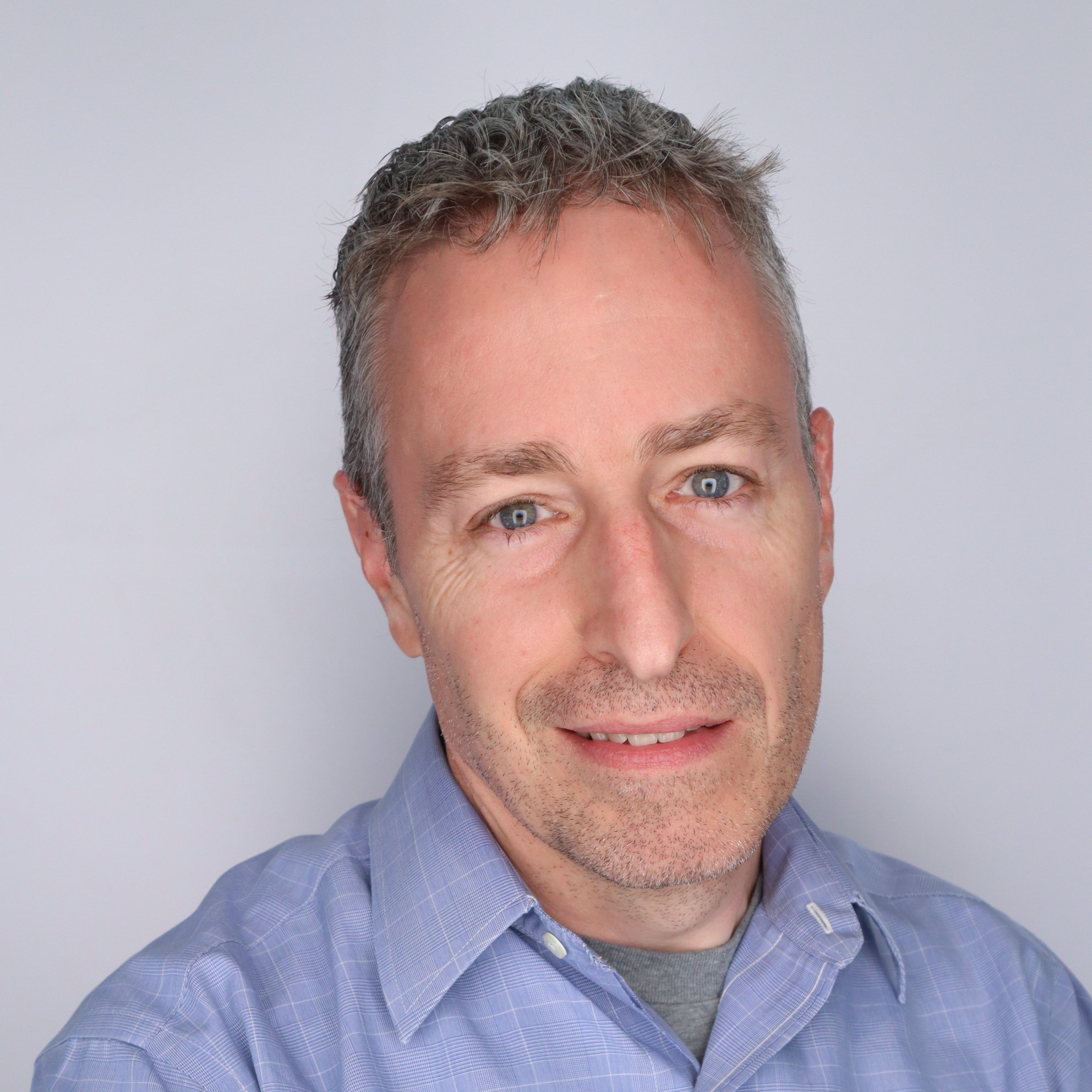 A close-up portrait of a middle-aged man with short, graying hair, blue eyes, and light facial stubble. He is wearing a light blue checkered collared shirt and smiling softly against a plain, light-colored background.