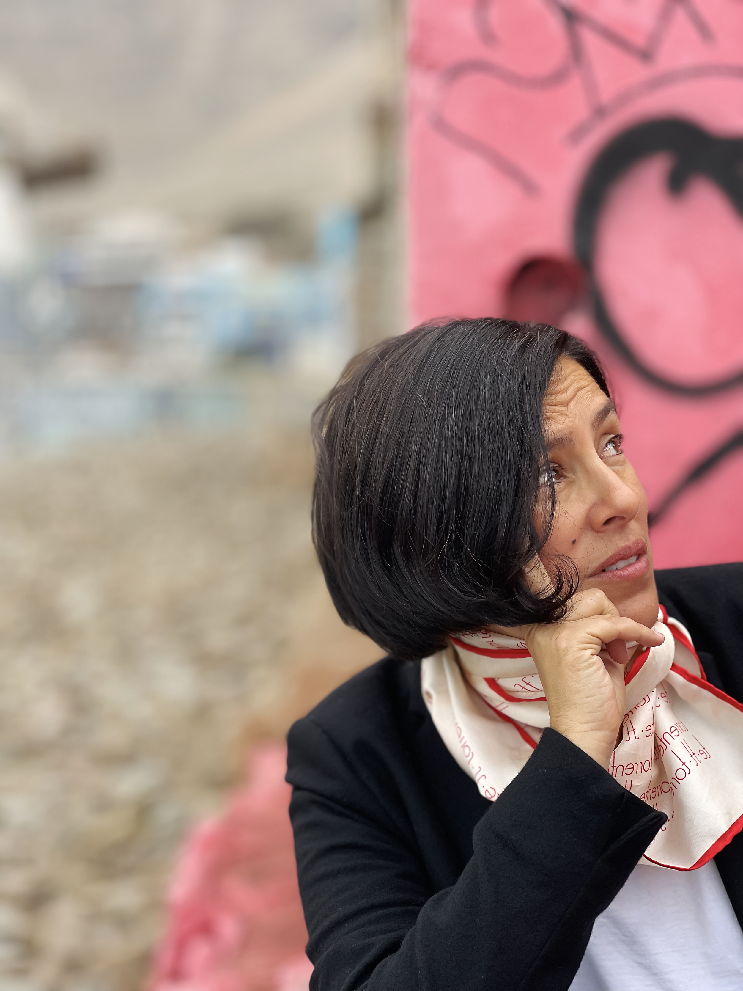 A woman with black, shoulder-length hair, wearing a black jacket and a white scarf with red writing, is looking to her right with a slight smile. The background is blurred with a pink graffiti wall on one side.