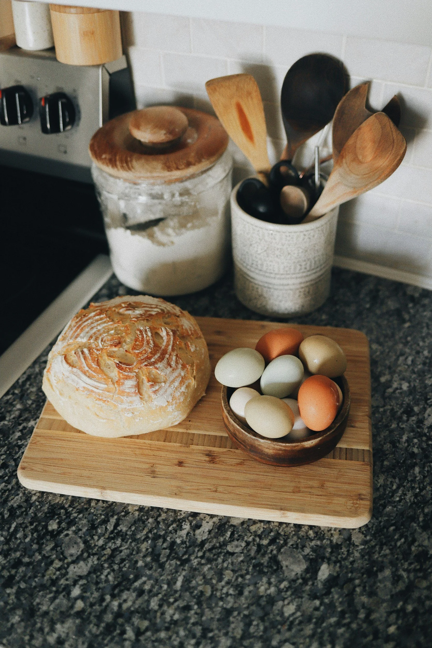 A kitchen countertop with a wooden cutting board holding a round loaf of bread and a small bowl of assorted eggs. In the background, there is a jar with a lid, a container with wooden spoons, and a stove with control dials.