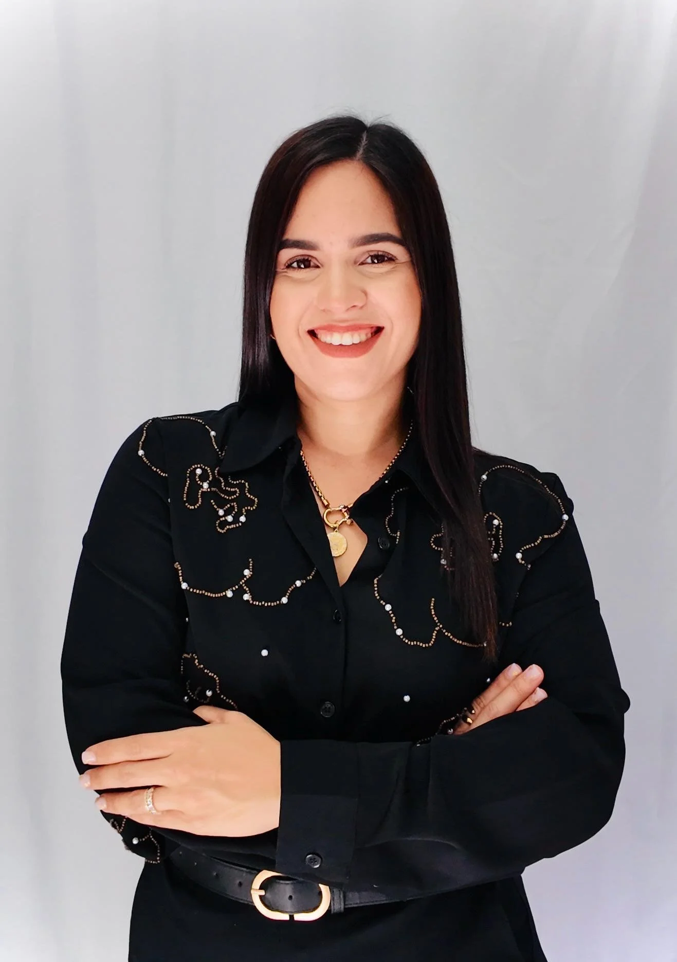 A woman with long dark hair smiling, wearing a black shirt adorned with pearl and chain embroidery, crossed arms, against a gray background.
