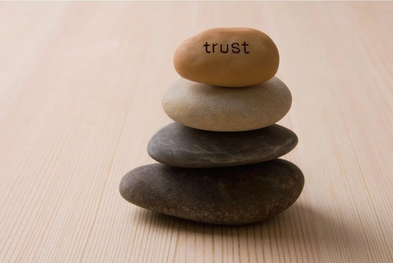 A stack of five smooth stones on a light-colored wooden surface, with the top stone having the word 'trust' written on it.