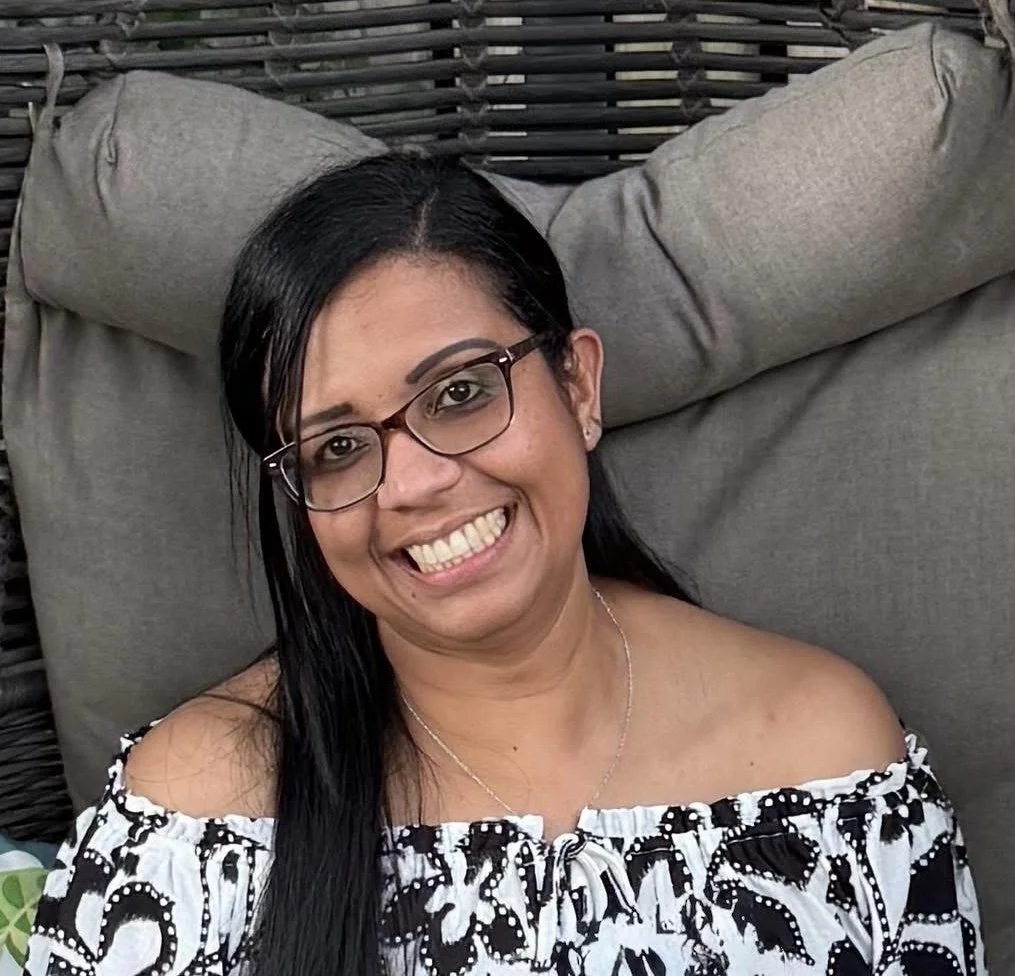 A woman with long black hair, glasses, and a big smile, wearing an off-shoulder black and white patterned top, sitting on a cushioned outdoor chair.