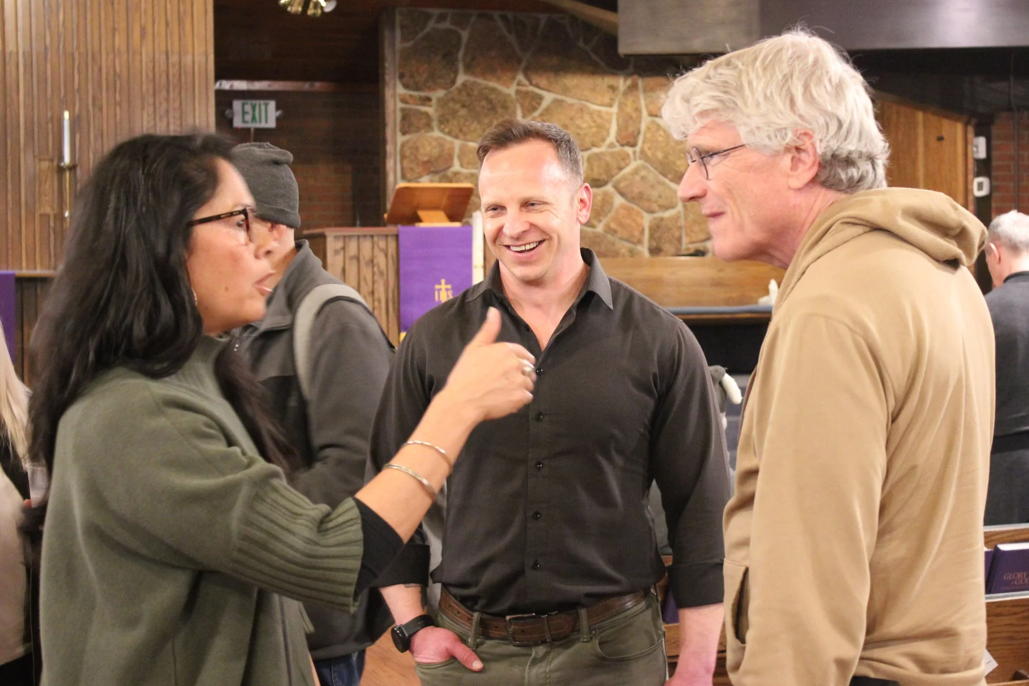 Four people engaged in conversation indoors, with a stone and wood background, and purple and gold decorations.