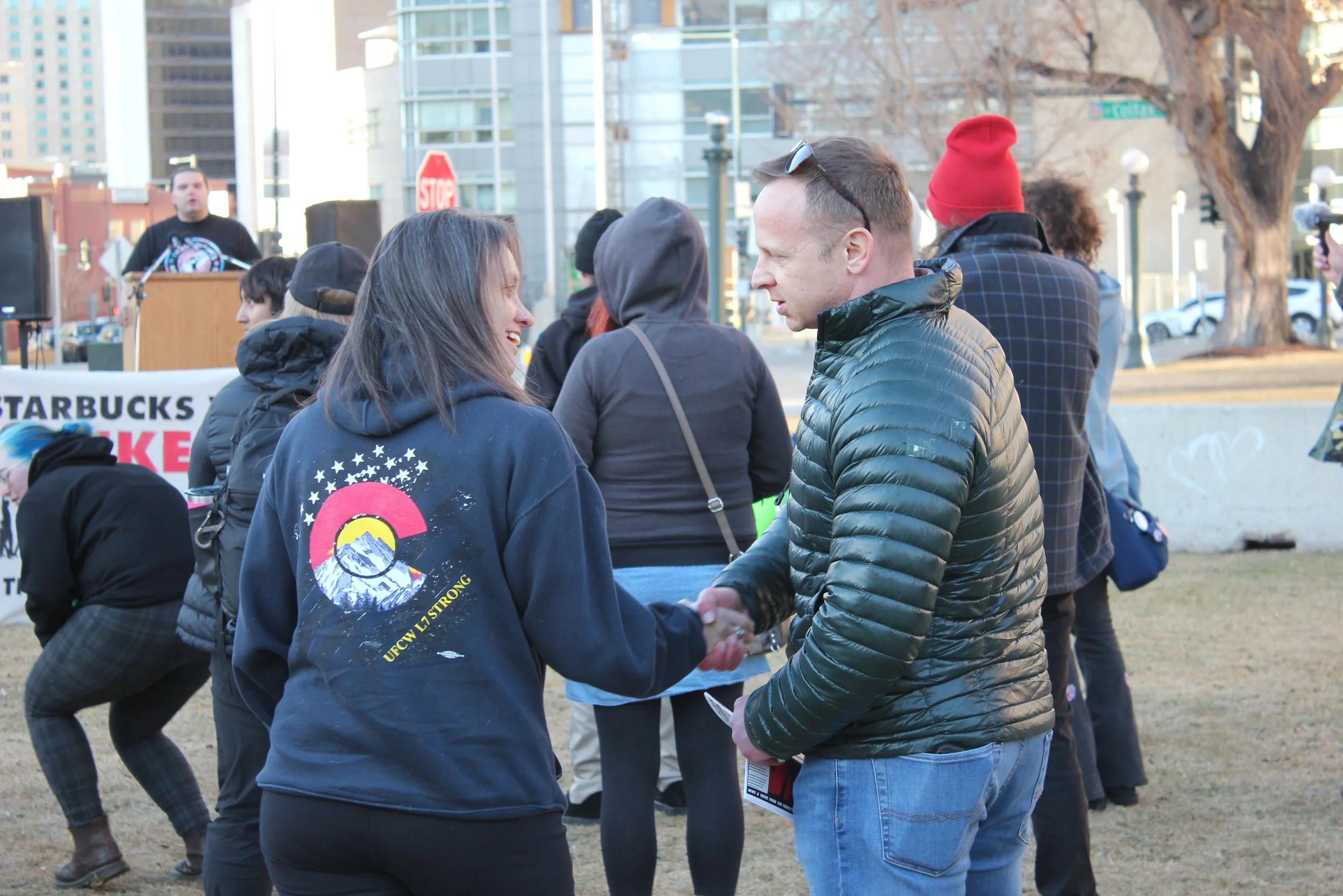 A woman and a man shake hands at an outdoor protest or rally. Other people are gathered around, some with backpacks, and a person is speaking at a podium in the background. There are buildings and a stop sign in an urban setting.