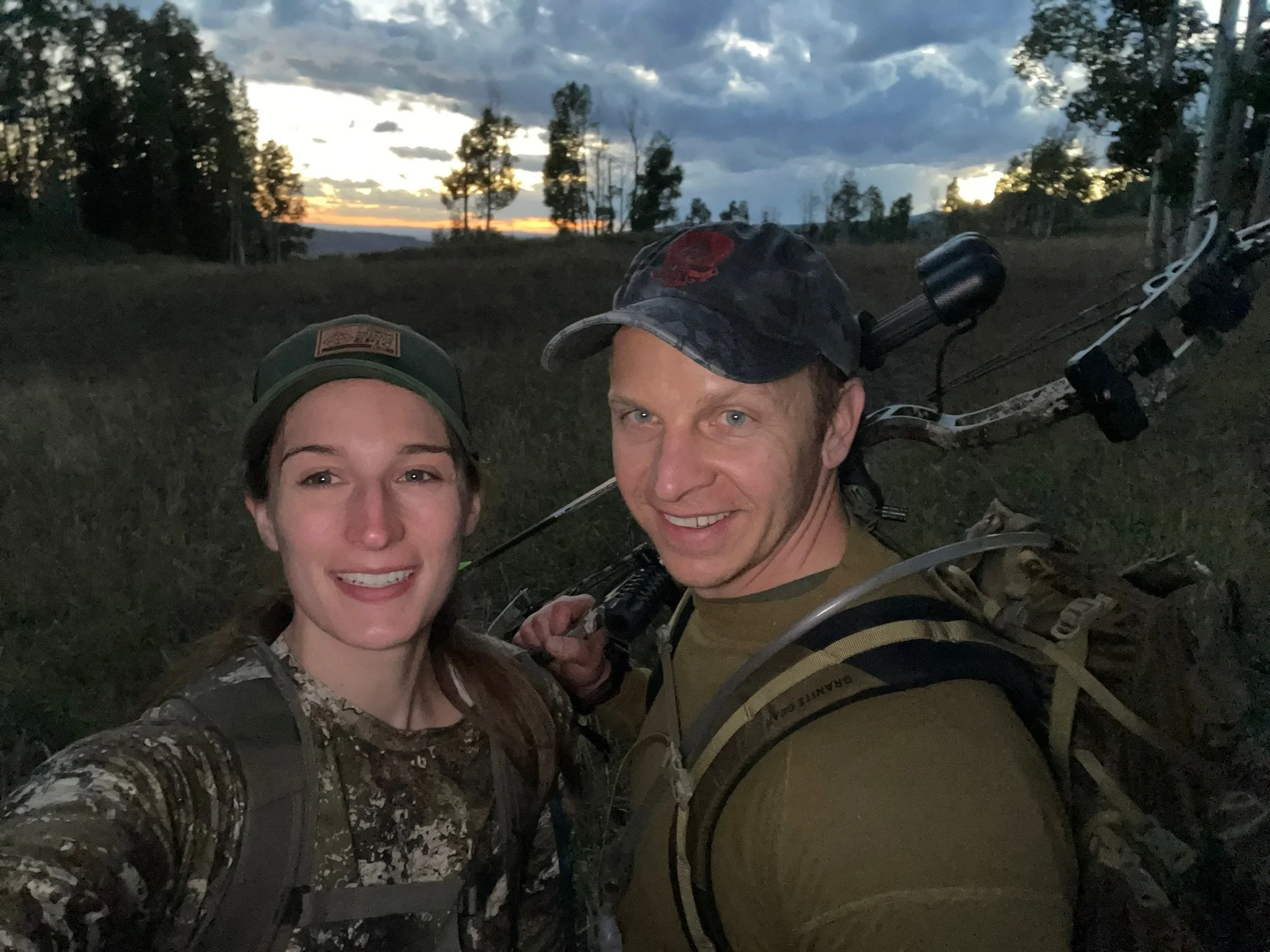 A smiling man and woman taking a selfie outdoors during sunset, carrying backpacks and a bow, with trees and a cloudy sky in the background.
