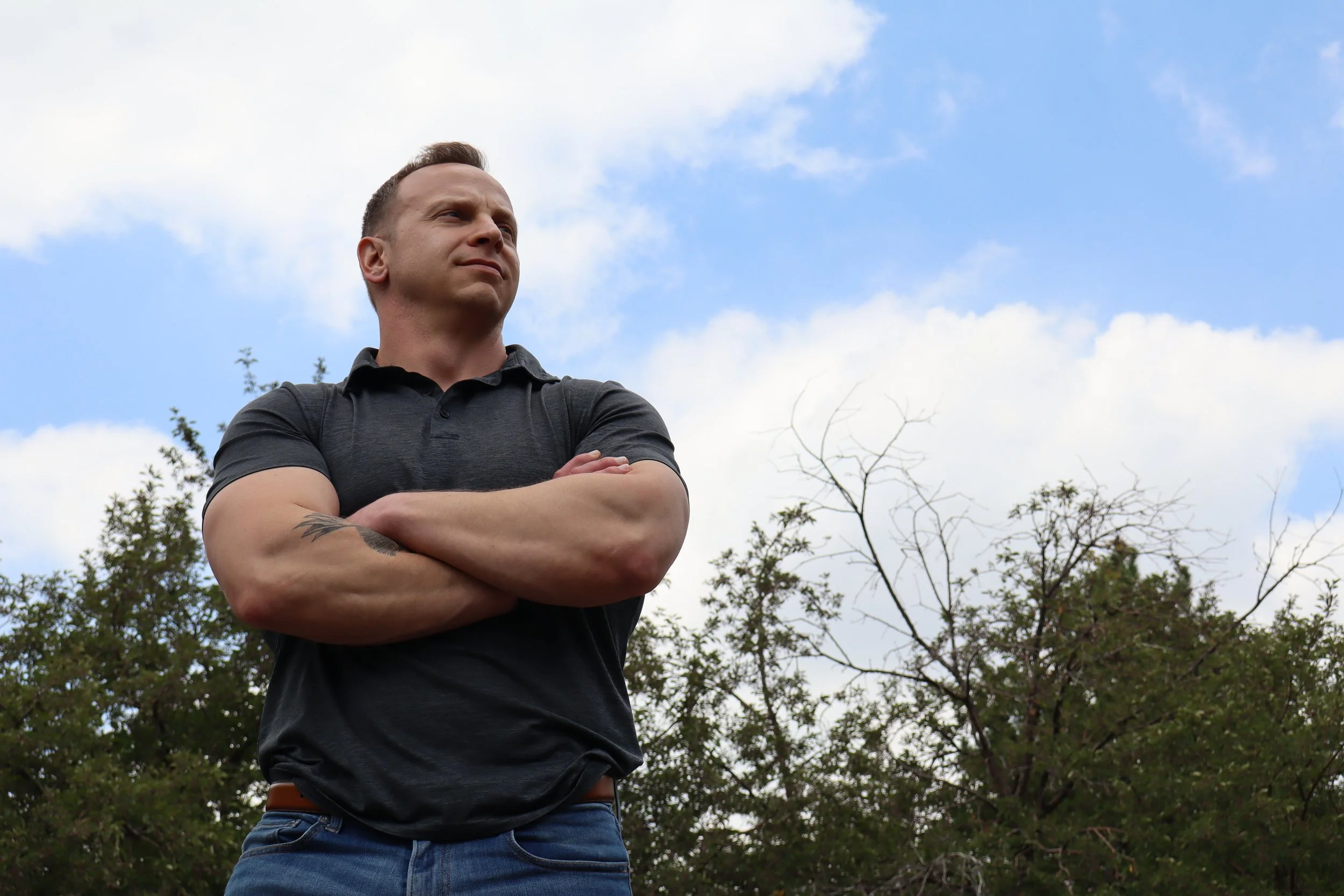 A man with short hair wearing a black polo shirt and blue jeans stands outdoors with his arms crossed, looking upwards. There are trees and a partly cloudy sky in the background.