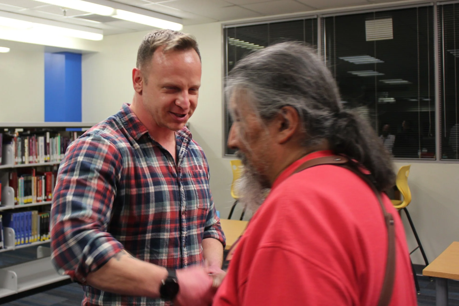 Two men shake hands and smile in a library or study room, with bookshelves and windows in the background.