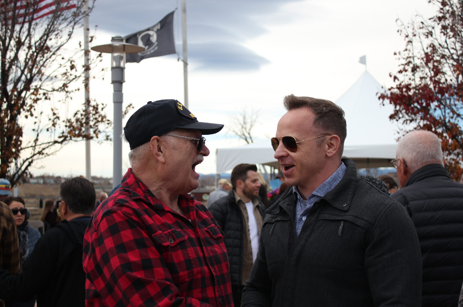 Two men talking and smiling outdoors at a social gathering, with a crowd, tents, and trees in the background.