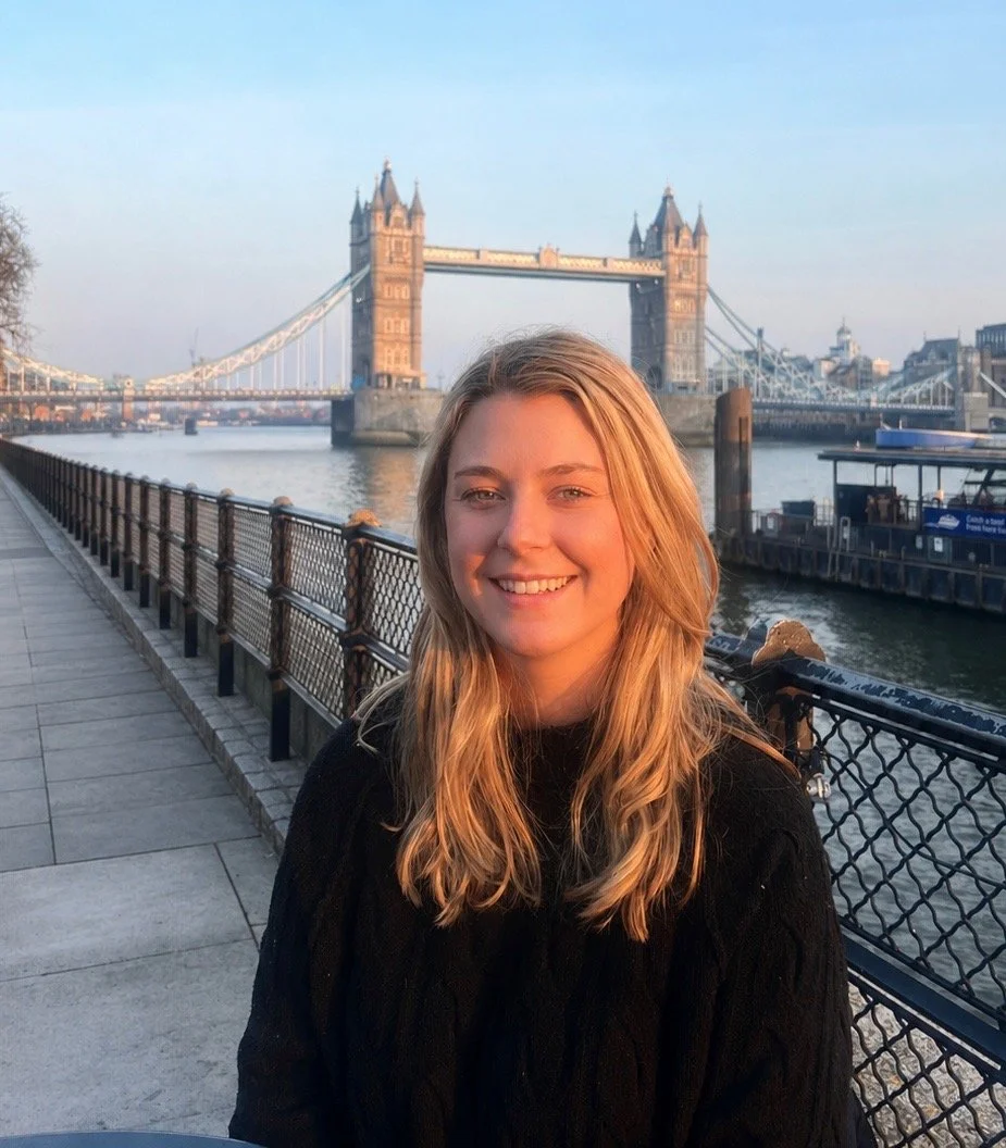 A young woman with long blonde hair smiling near the River Thames with Tower Bridge in the background in London, England, during sunset.
