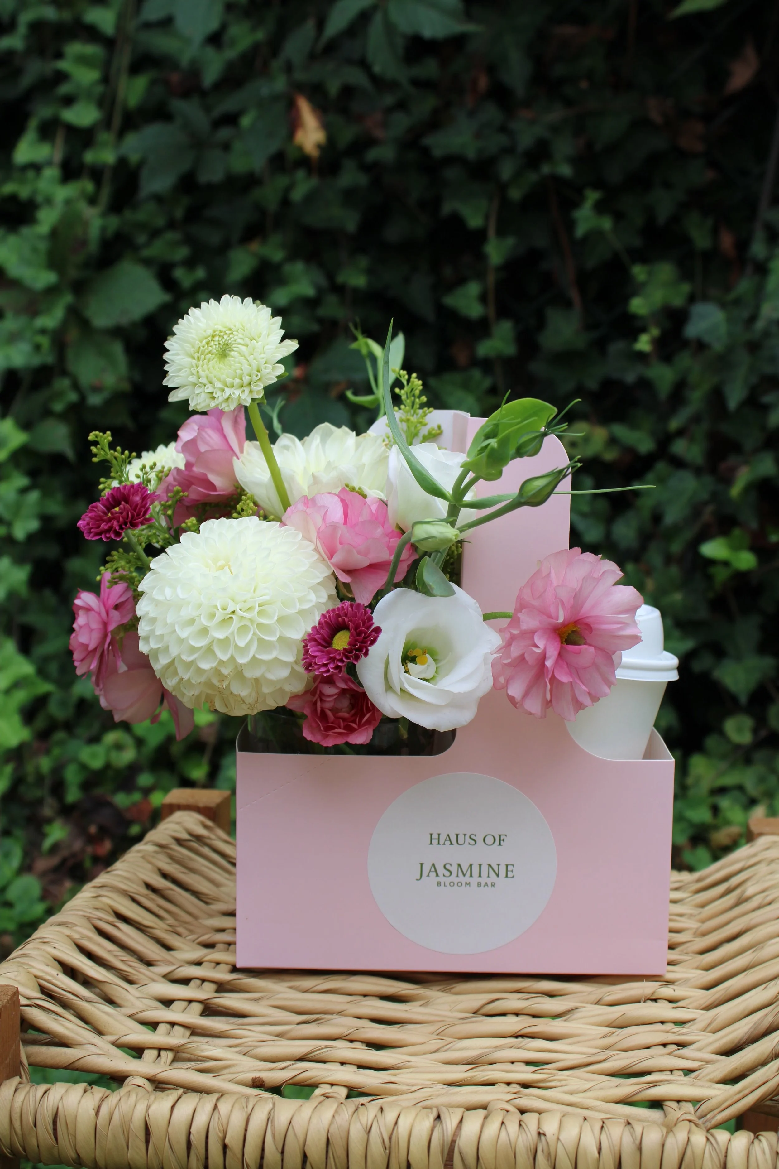 A bouquet of pink, white, and purple flowers in a pink paper bag labeled "Haus of Jasmine Flower Bar" placed on a woven wicker basket with a green leafy background.