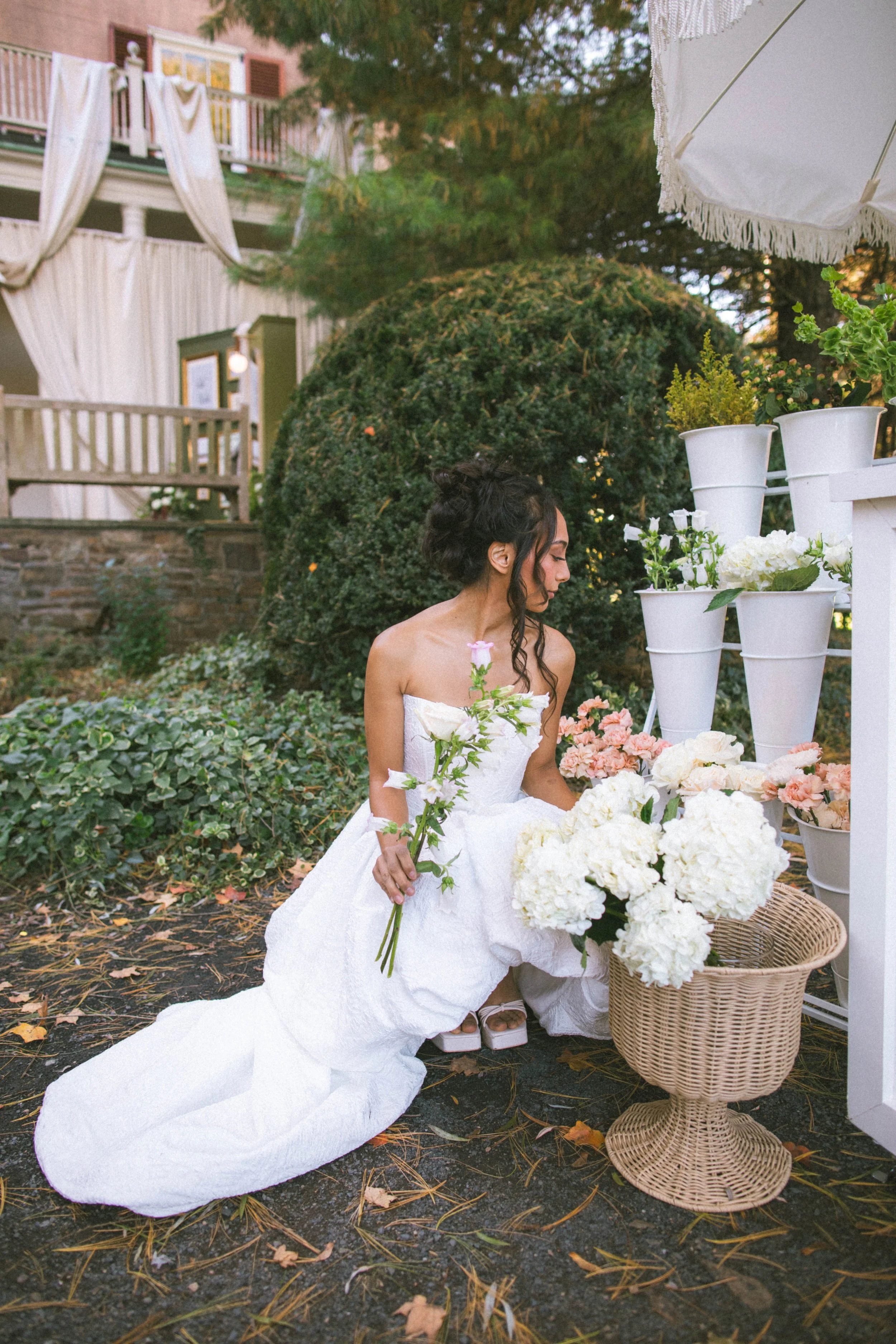 A young woman in a white wedding dress kneeling outdoors, holding a flower, surrounded by large bouquets of white and pink flowers, with a basket nearby. There is greenery, a bush, and a building with balconies and curtains in the background.