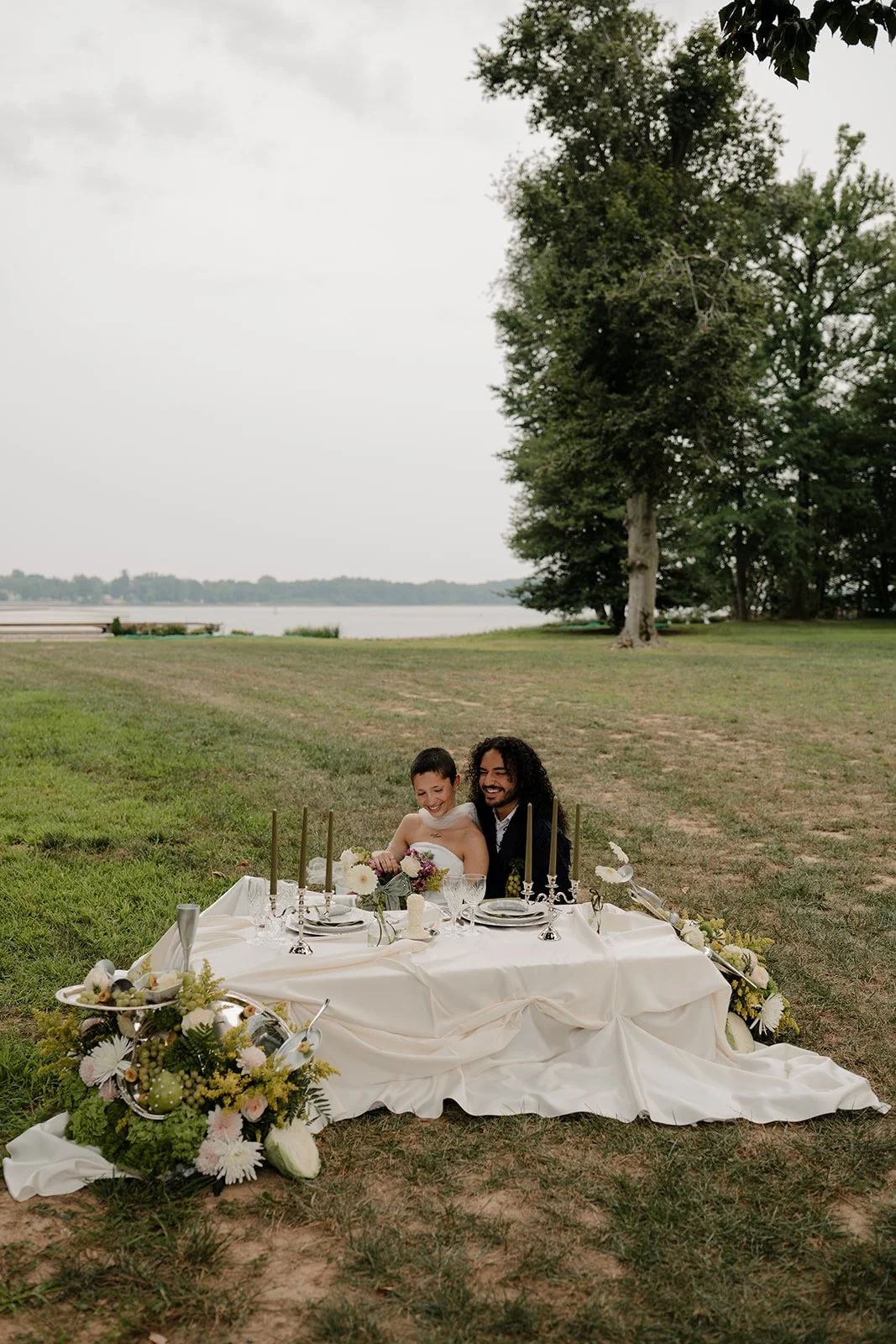 A couple, a man and a woman, sitting at a decorated outdoor table near a lake, smiling and enjoying each other's company, with trees and water in the background.