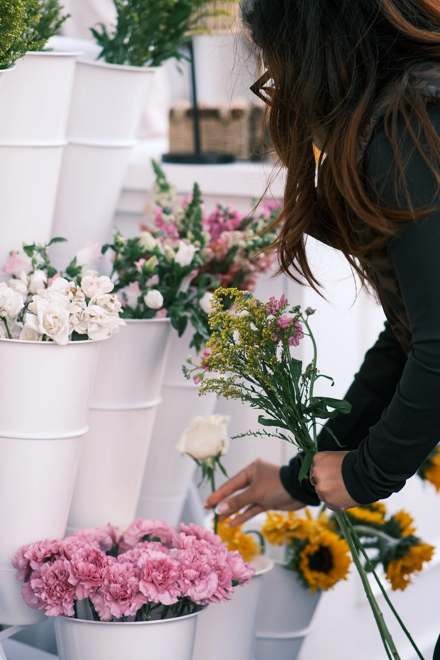 A woman arranging pink, yellow, and white flowers in white buckets at a flower shop.
