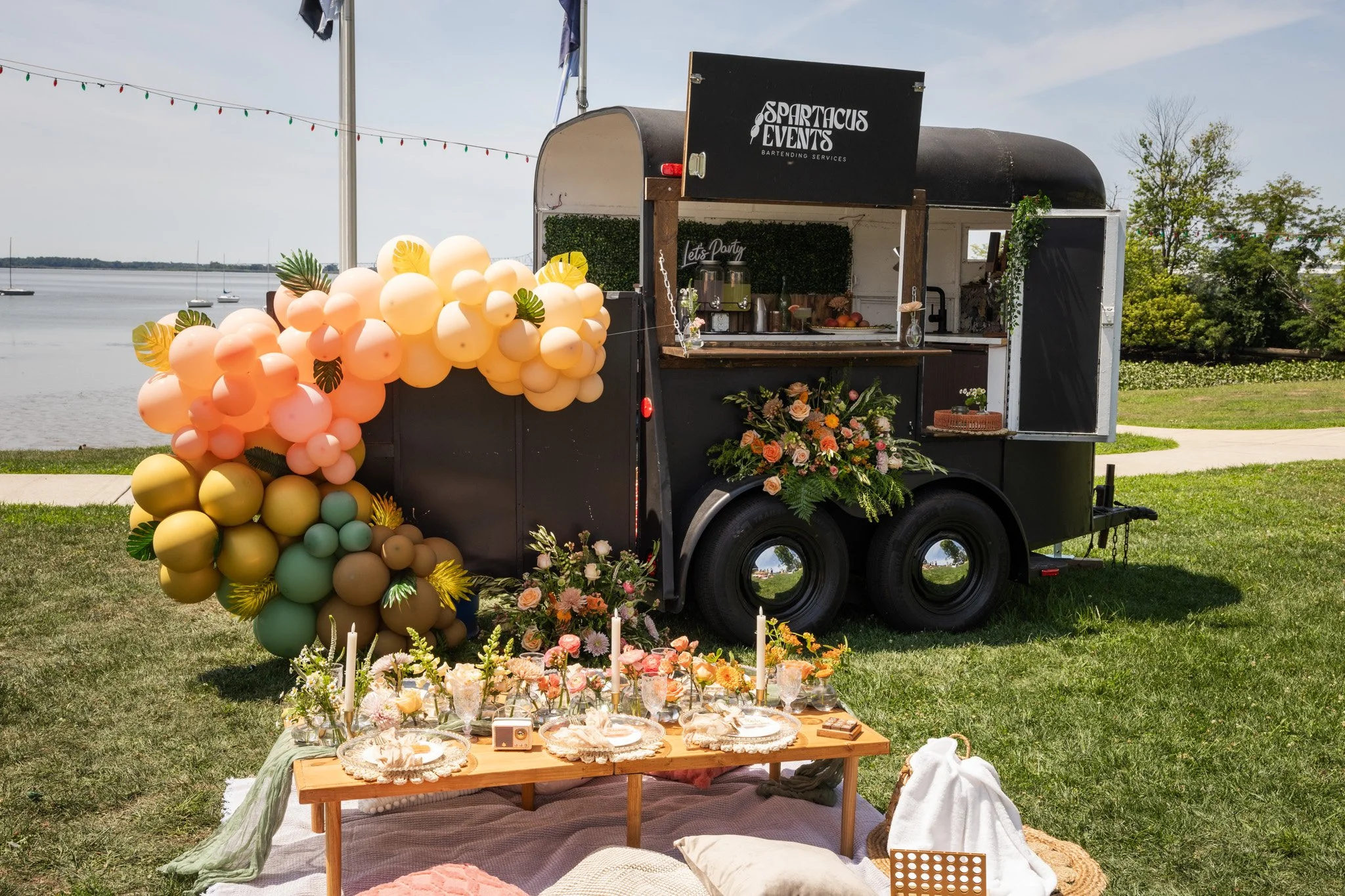 A mobile bar setup named Sparta Cup Events with balloons, floral arrangements, candles, and tableware outdoors by a lake, with sailboats and trees in the background.
