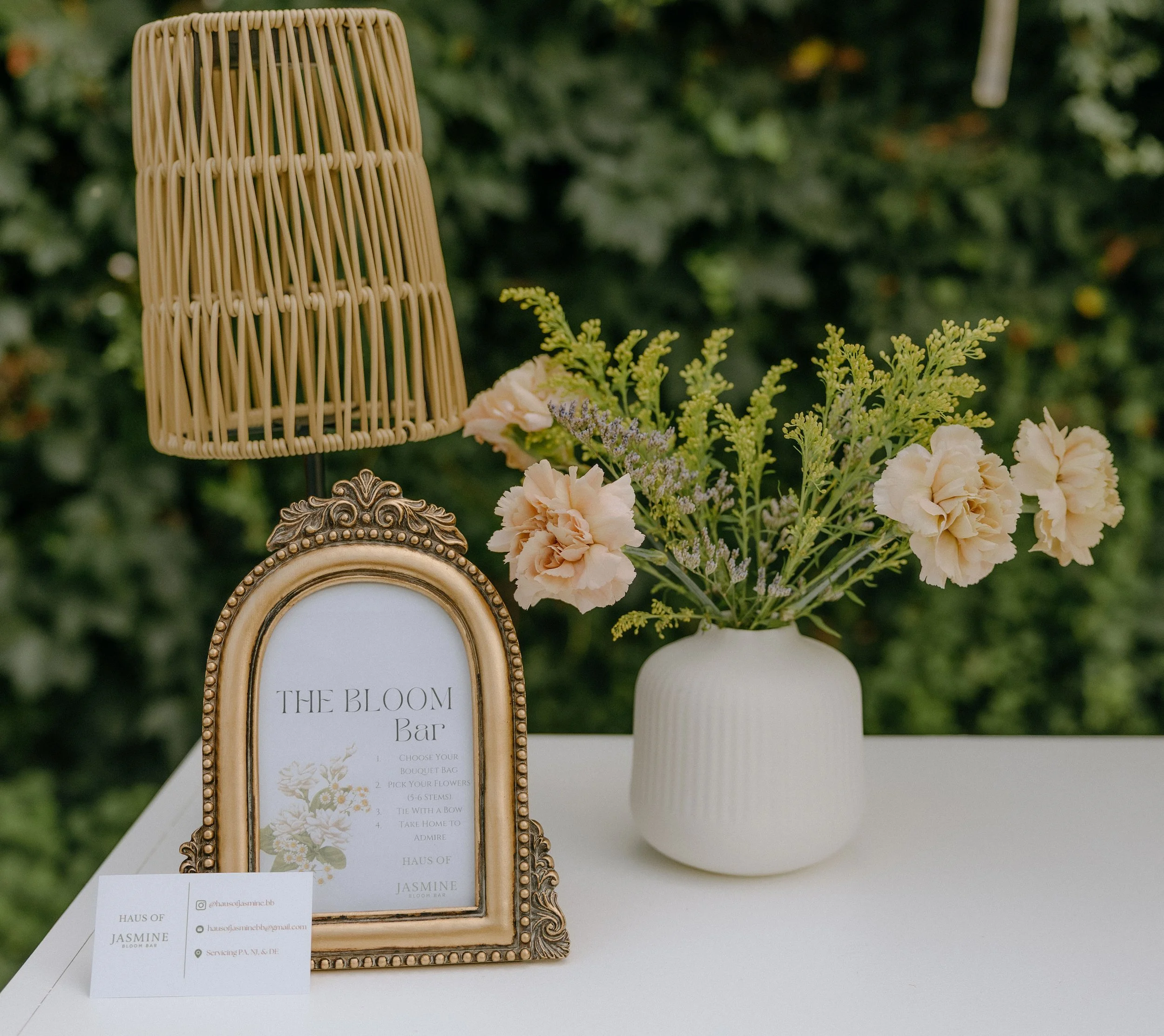 A decorative table display featuring a gold ornate picture frame with a sign inside titled 'The Bloom Bar' and instructions for a floral bouquet, a white vase holding a mixed arrangement of light pink and white flowers with green foliage, and a tan-colored wicker lampshade in an outdoor setting with greenery in the background.