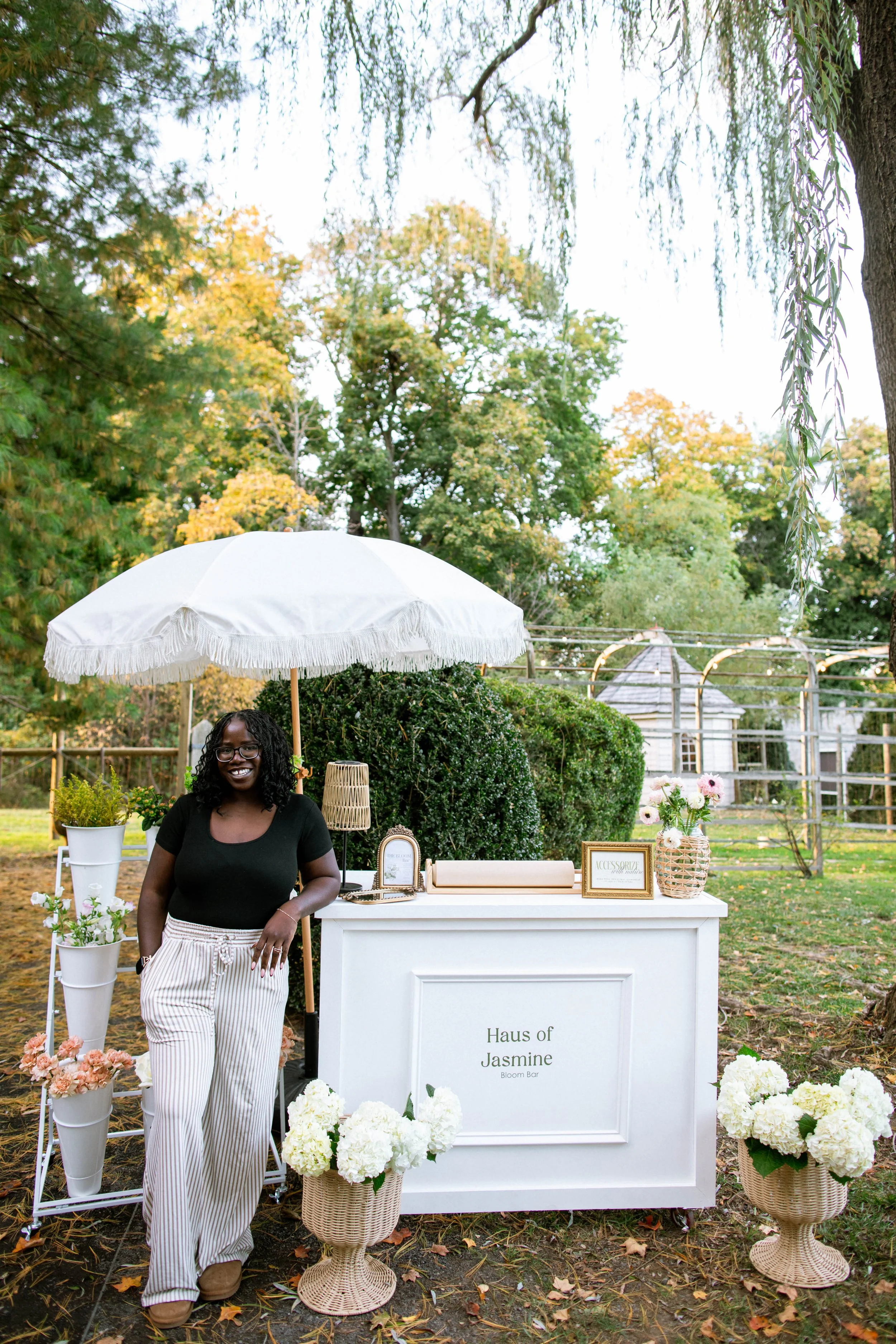 A woman standing in front of a white desk at an outdoor event, with flowers and decorative items displayed on and around the desk, under a large white umbrella. There are trees with green and yellow leaves in the background.