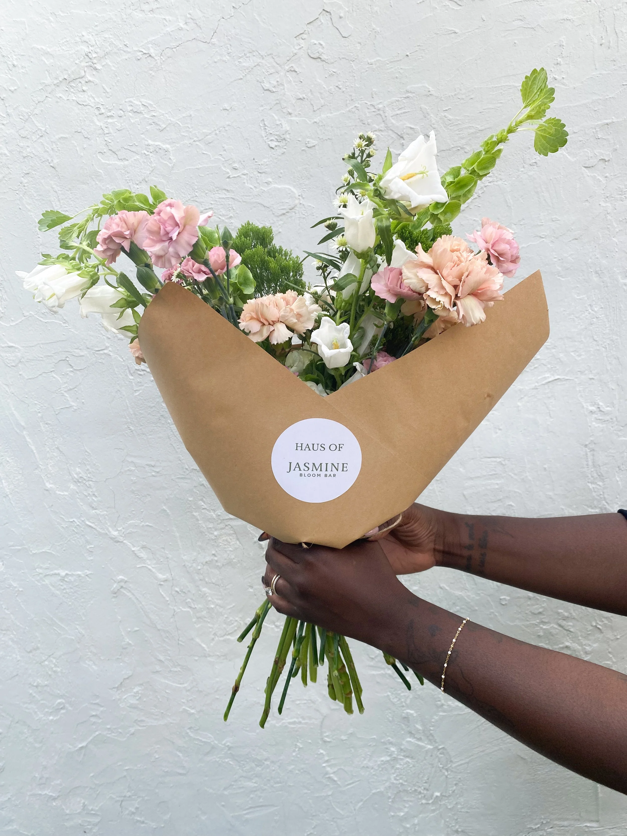 Person holding a bouquet of pink, white, and green flowers wrapped in brown paper against a textured white wall.