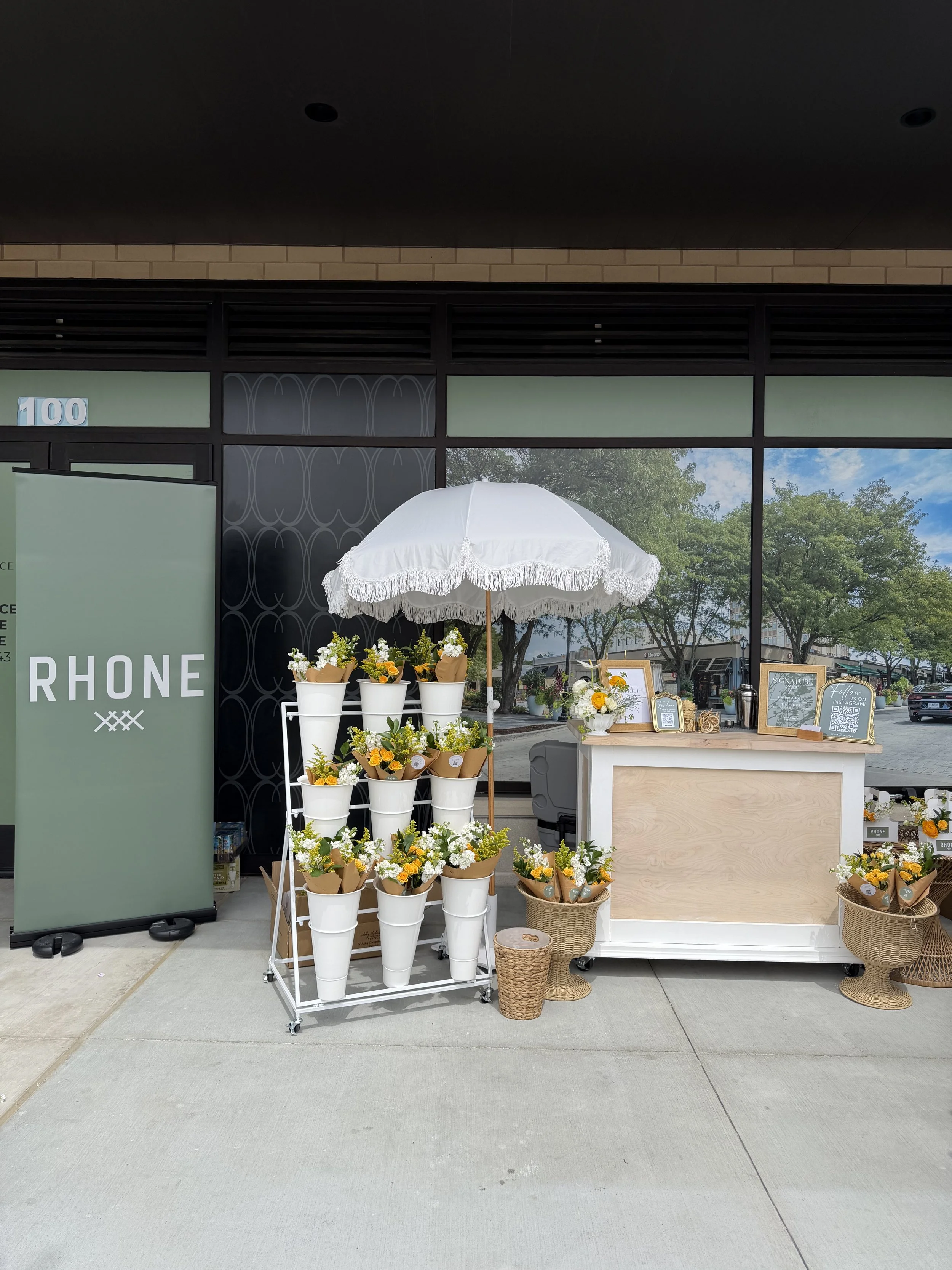 Flower display stand with white buckets filled with yellow and white flowers, a white parasol, and framed signs on a light wood counter outside a building with large glass windows and brick accents.