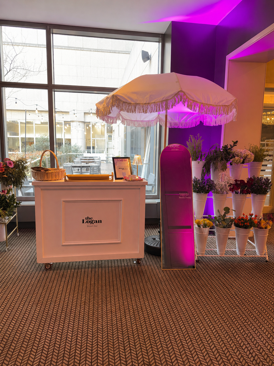Indoor floral display station with a white table labeled 'the Logan Bloom Bar', a large white parasol with fringed edges under purple lighting, and various potted flowers on white shelves and on the floor, with large window and outdoor patio visible in the background.