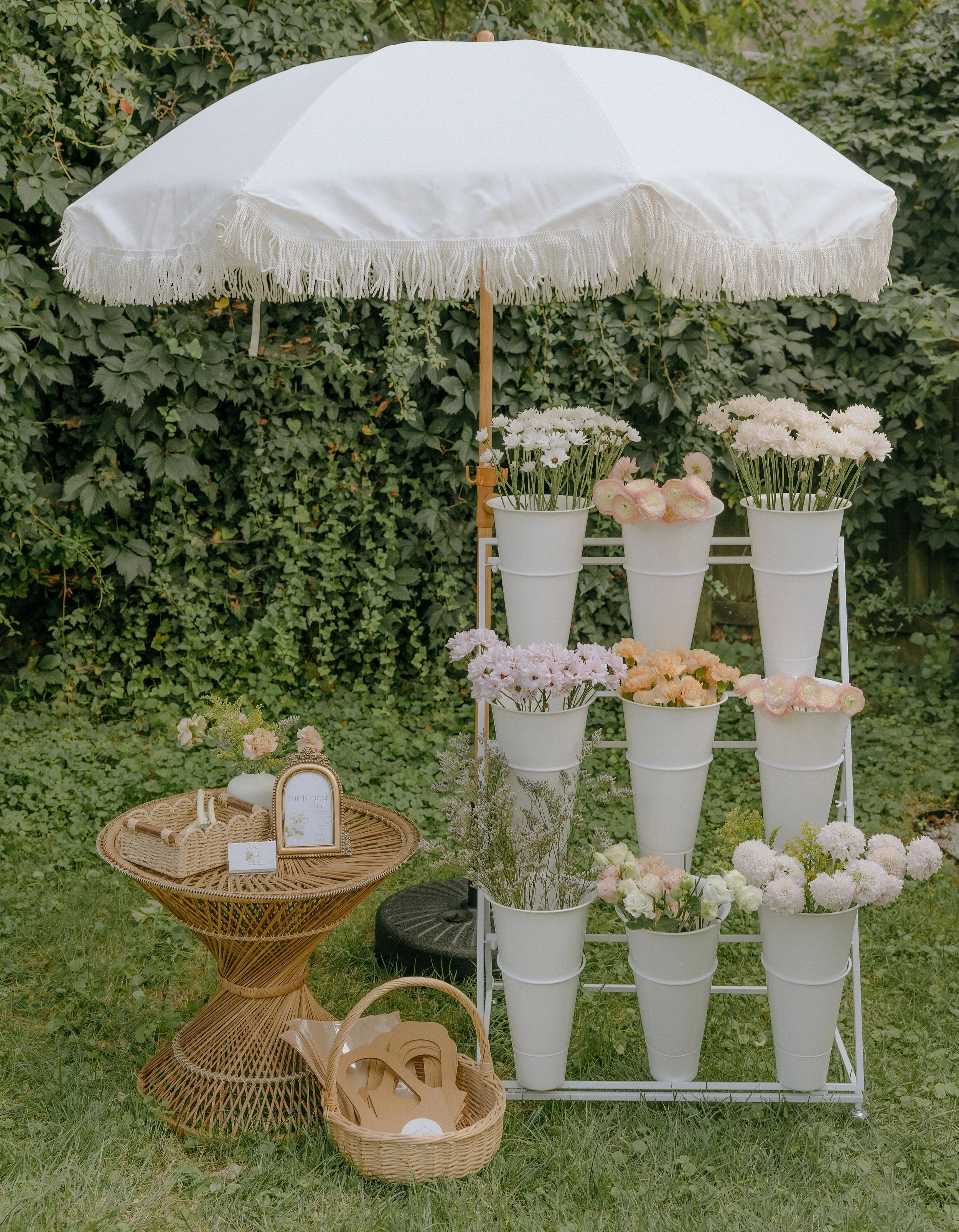 An outdoor floral display with white flowers in white buckets arranged on a white rack, a white fringe umbrella shading the display, and a wicker table with small decorative items and signs, set against a background of green foliage.