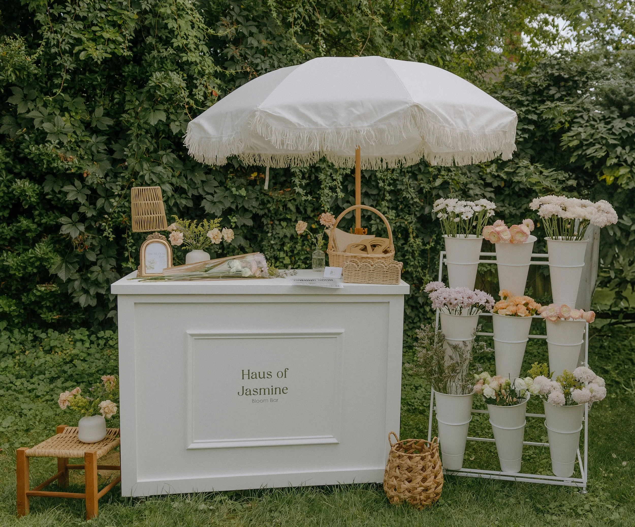 Outdoor display of flowers, with a white umbrella shading a table labeled "Haus of Jasmine Bloom Bar," decorated with flowers, baskets, and small decorative items, against a background of green foliage.