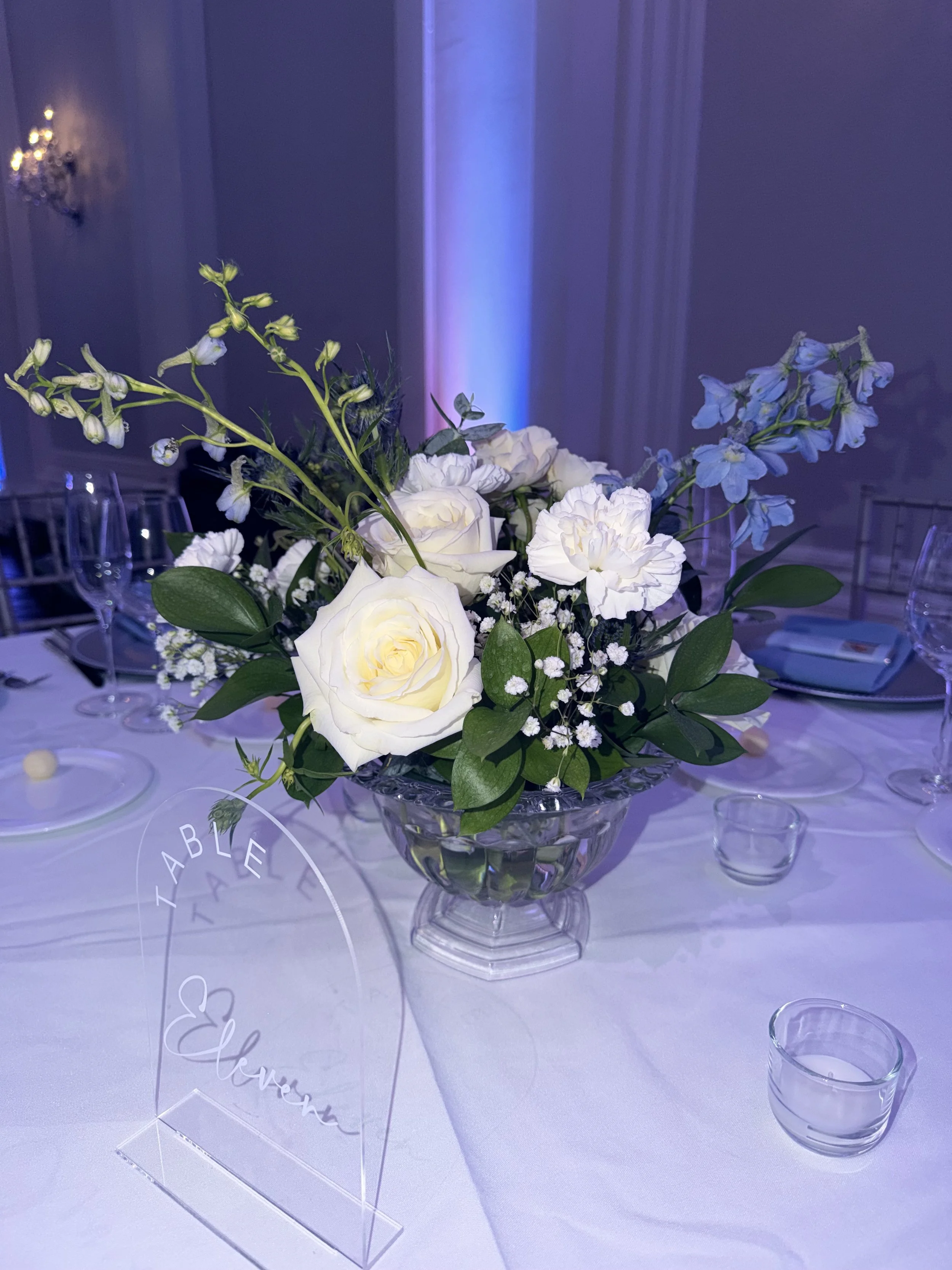 A floral centerpiece with white roses, blue delphiniums, and green leaves in a glass vase on a white table, surrounded by glass candle holders and clear acrylic signs with white cursive writing.