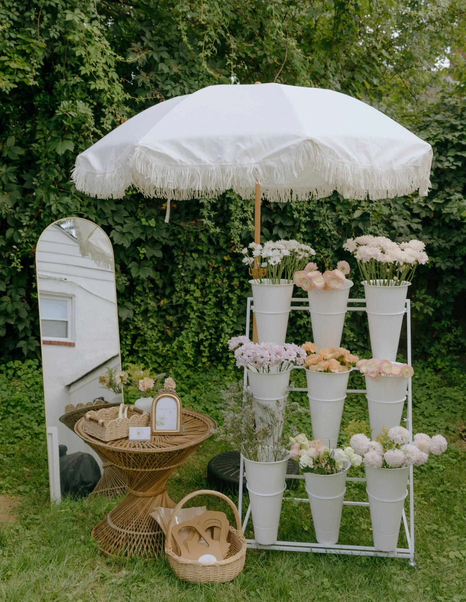 A white outdoor parasol with fringe, surrounded by white flower pots filled with pink and white flowers, set against a green leafy background. There is a white mirror and a wicker table with small decorative items nearby.