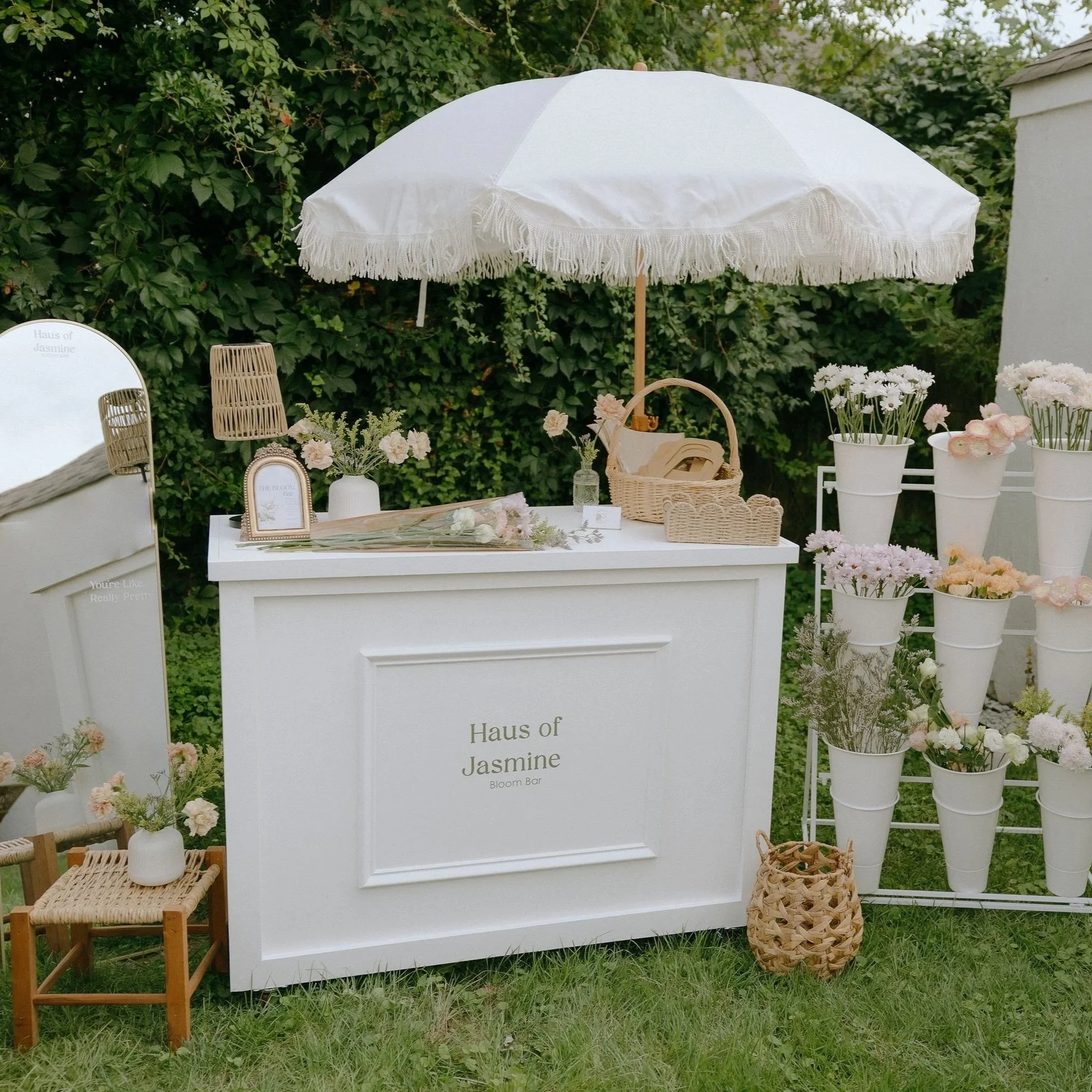 Outdoor setup with a white sign that says "Haus of Jasmine Bloom Bar" surrounded by white and pink flowers in vases, wicker and wooden furniture, and a large white parasol against a backdrop of green foliage.