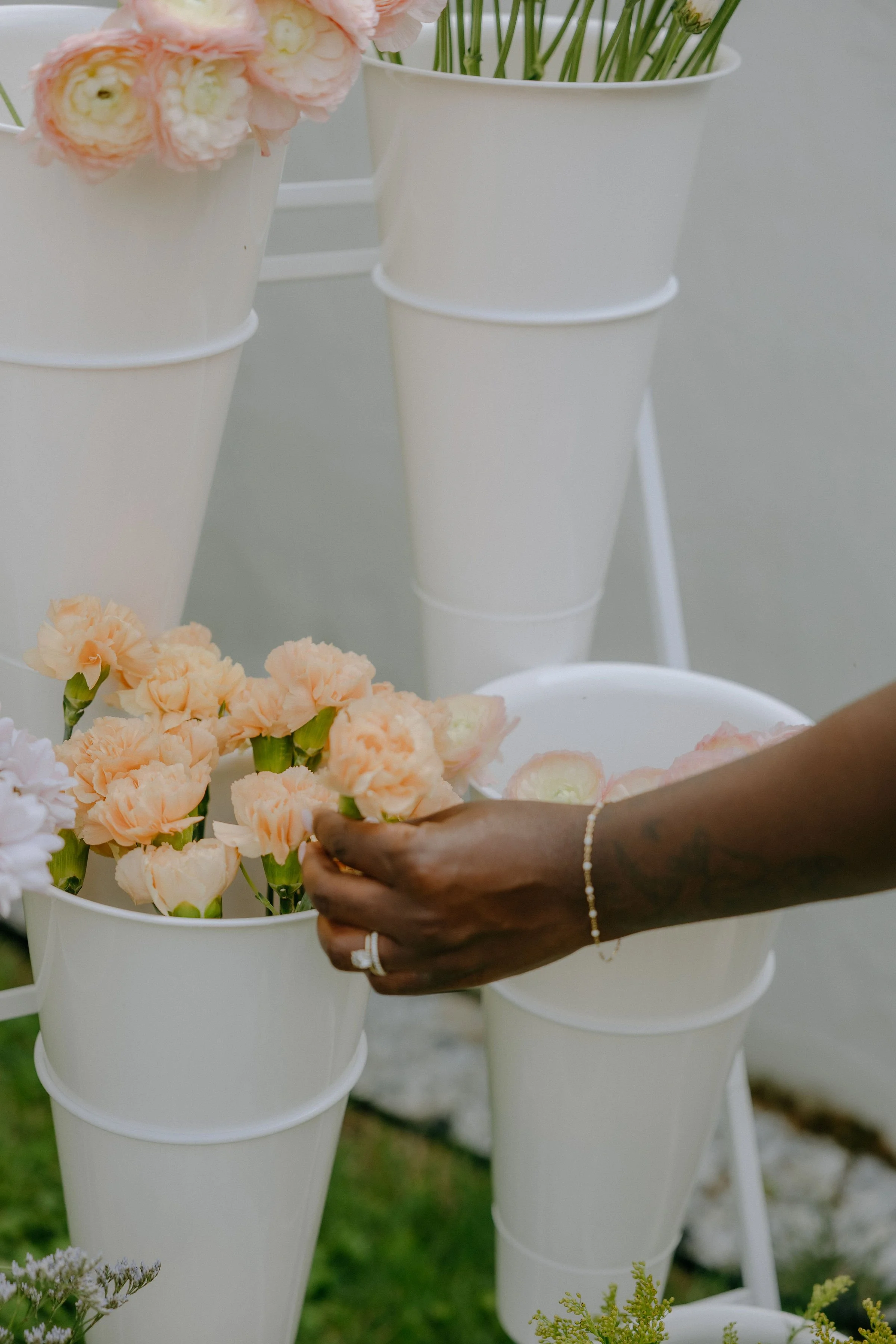 A person's hand is arranging a pale orange flower in a white bucket, with more flowers in other buckets in the background.