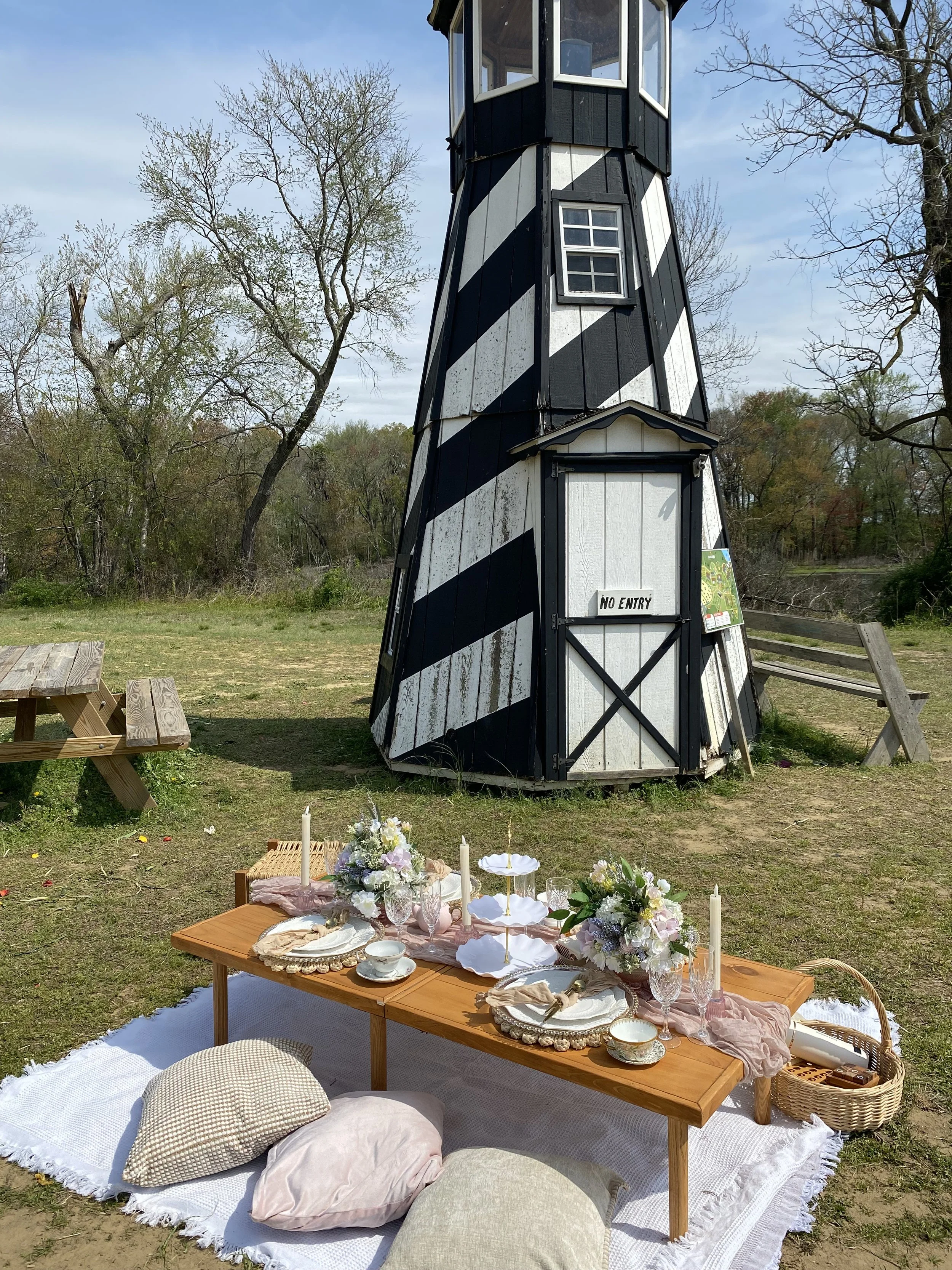 An outdoor picnic setup with a low wooden table decorated with flowers, candles, and fine china, on a white blanket, in front of a black and white striped lighthouse surrounded by trees.