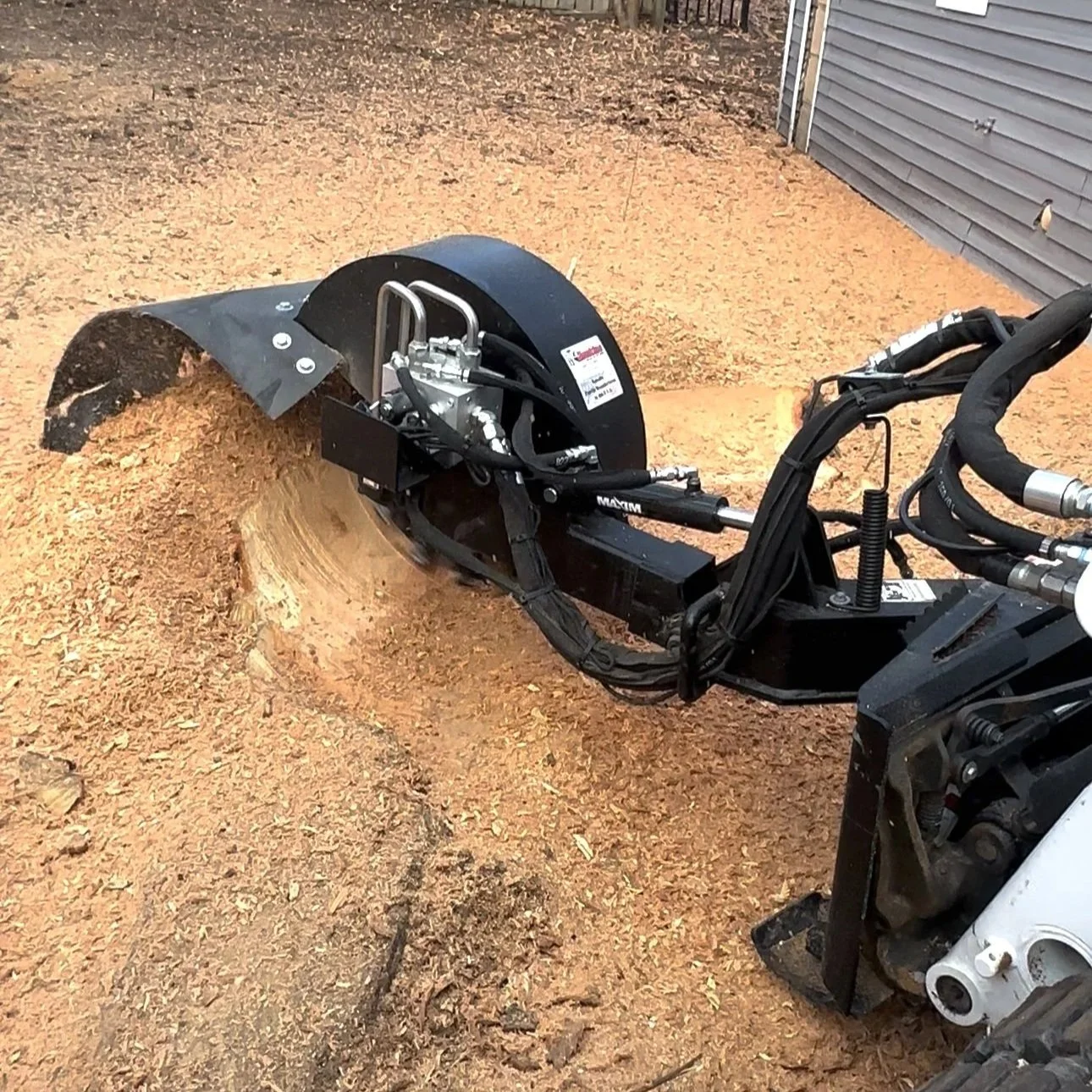 Bobcat stump grinding machine in use, processing stump on the ground next to a building with gray siding.