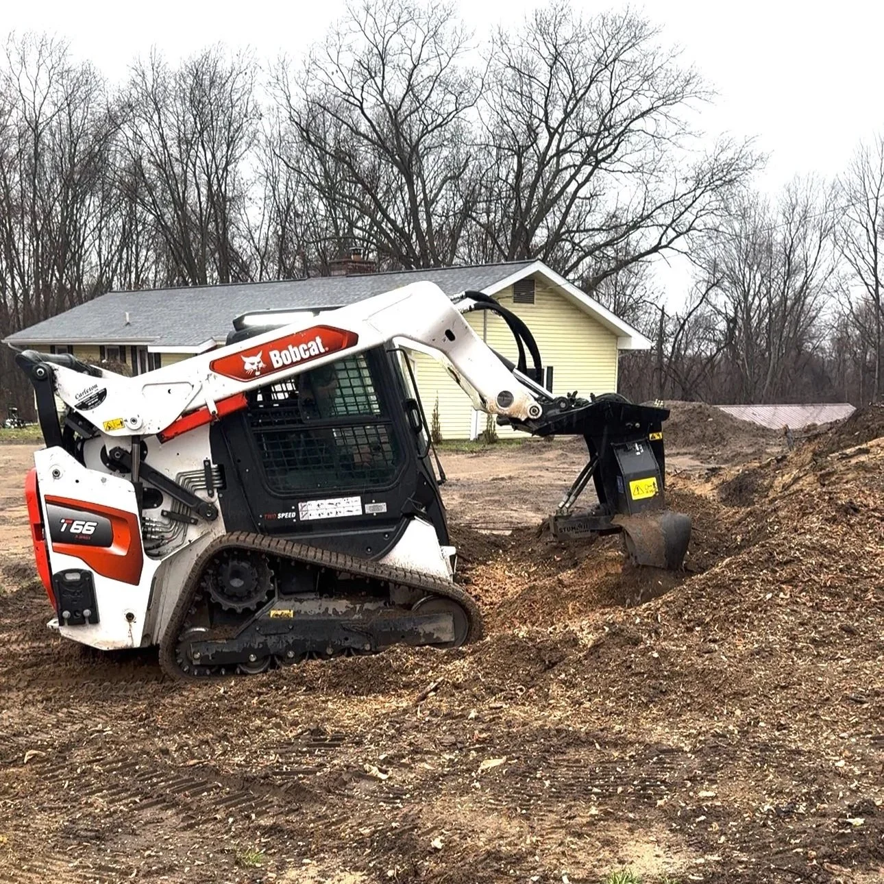 A Bobcat T66 compact track loader working on a construction site with a house and leafless trees in the background. Bobcat is stump grinding